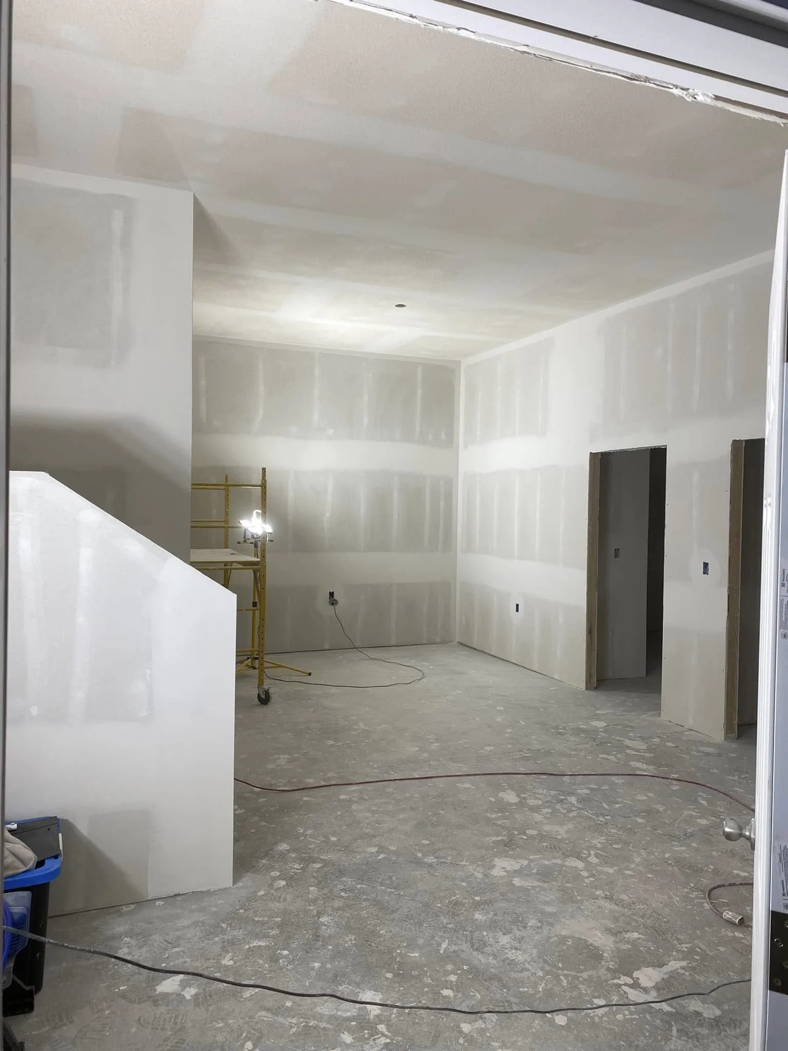 Interior of a room under construction with drywall installed on the walls and ceiling, unfinished flooring, and construction equipment including a yellow scaffold and electrical outlets.