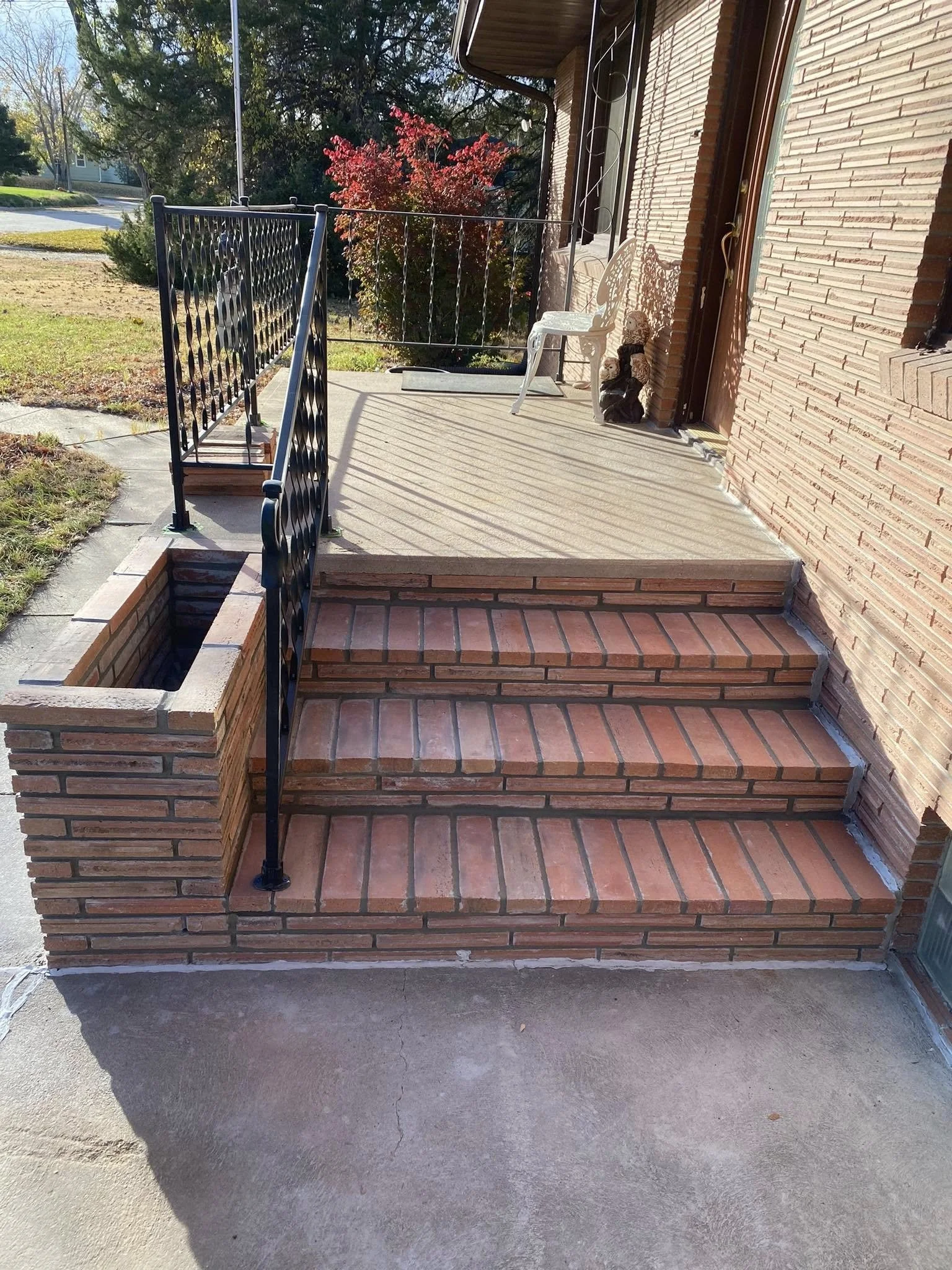 Front porch with brick steps, black metal railing, white chair, and decorative garden gnome at the entrance of a house. Fallen leaves on the porch, bright sunlight, trees, and a red bush in the background.