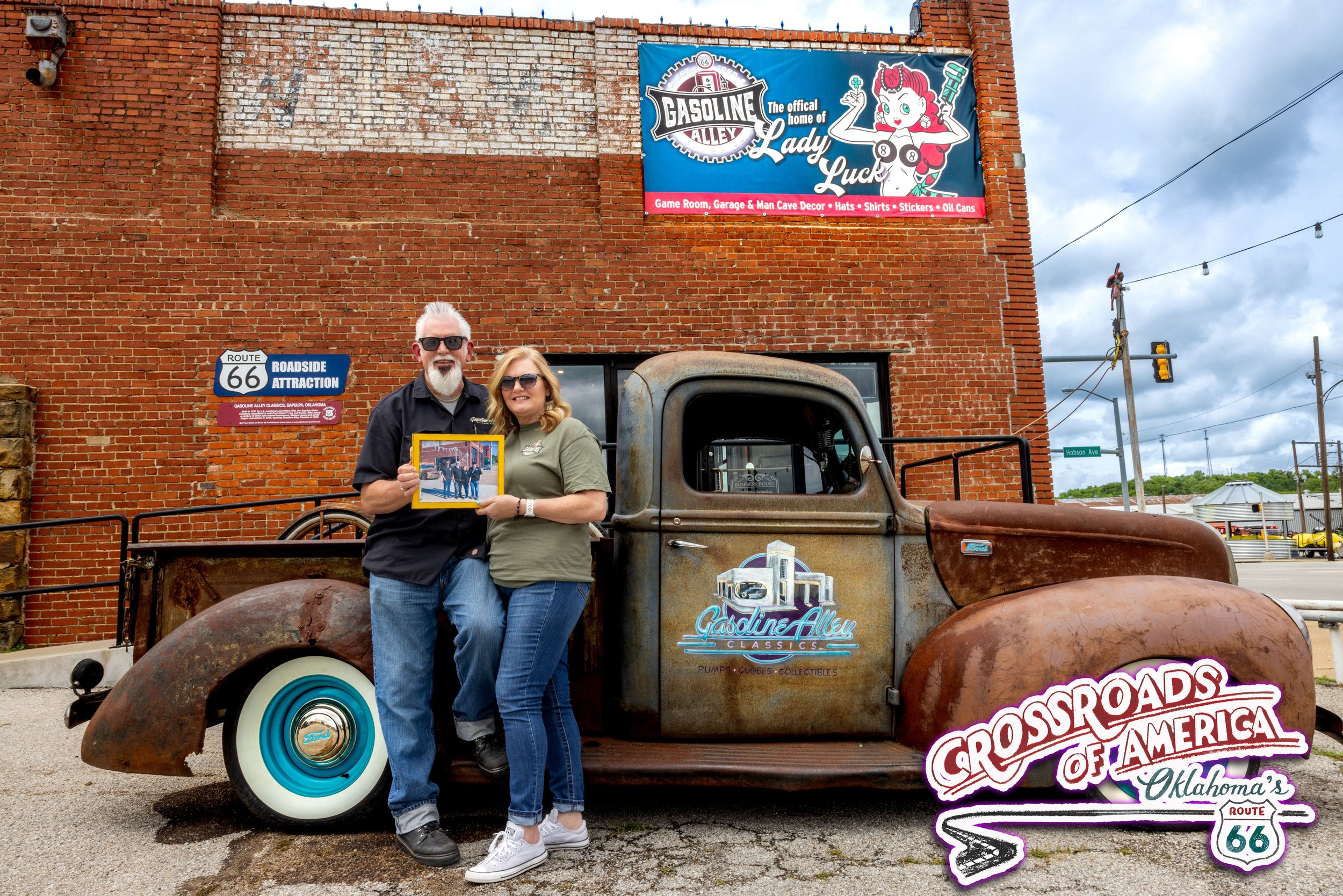 A man with white hair, beard, and sunglasses standing next to a woman with blonde hair and sunglasses, holding a framed photo, in front of an old rusty truck with gasoline alley signage. They are outside on a street with brick buildings and Route 66 signs in the background.