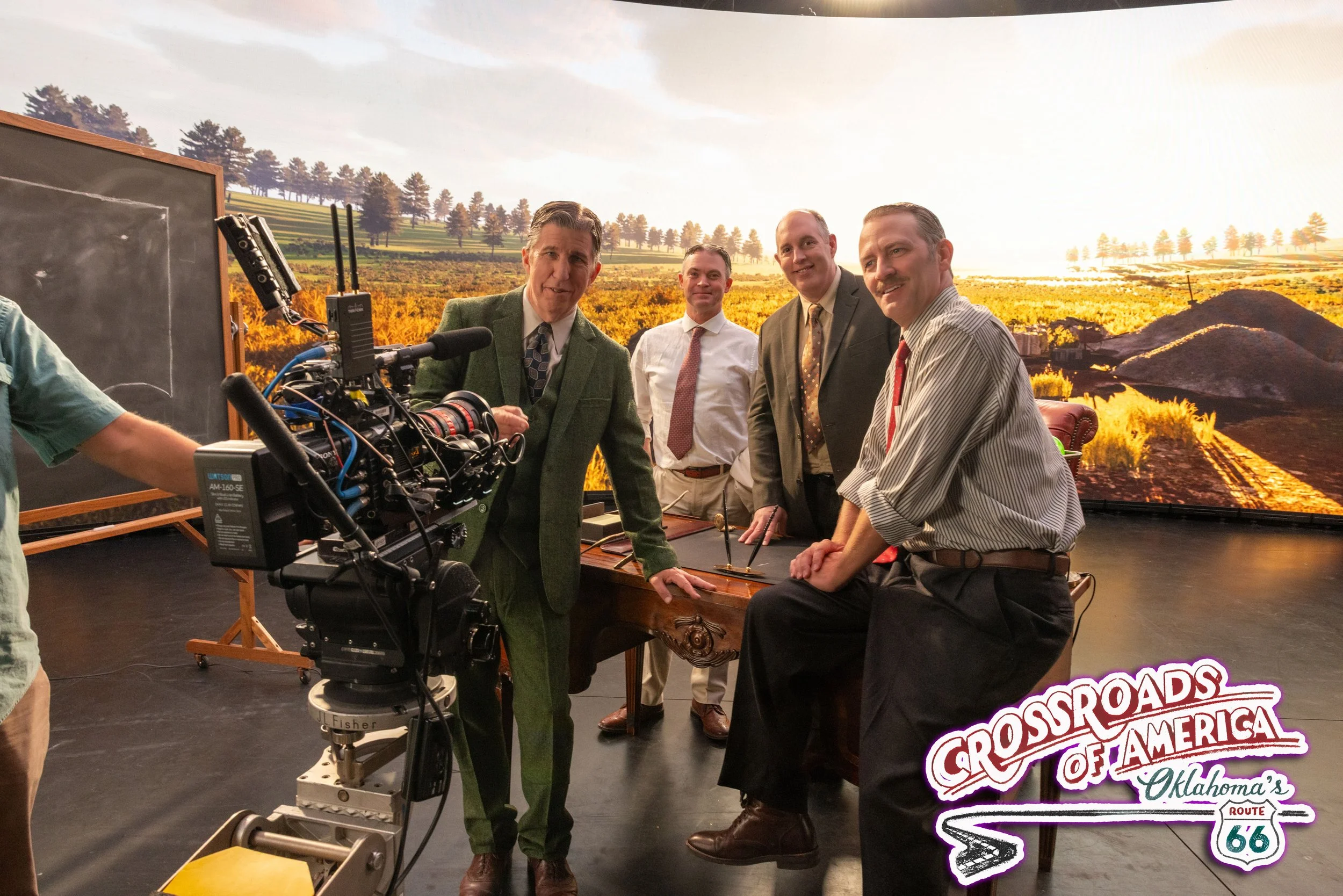 Four men in business attire standing behind a desk with a camera pointing at them; background features a large scenic wall mural of a rural landscape with trees and fields, and a logo reading 'Crossroads of America Oklahoma's Route 66.'