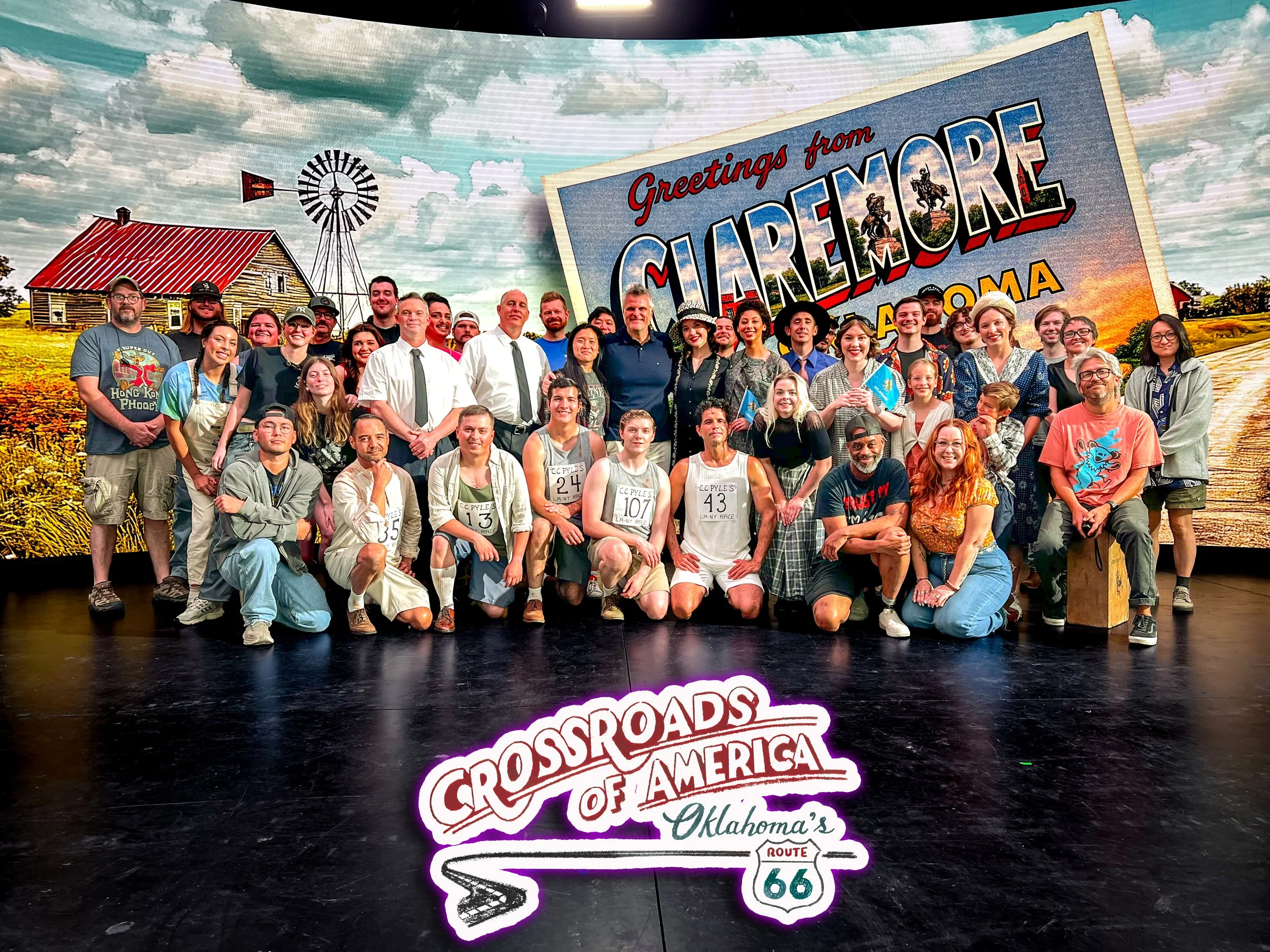 Group of cast and crew standing on a sound stage, and a logo reading 'Crossroads of America Oklahoma's Route 66.'