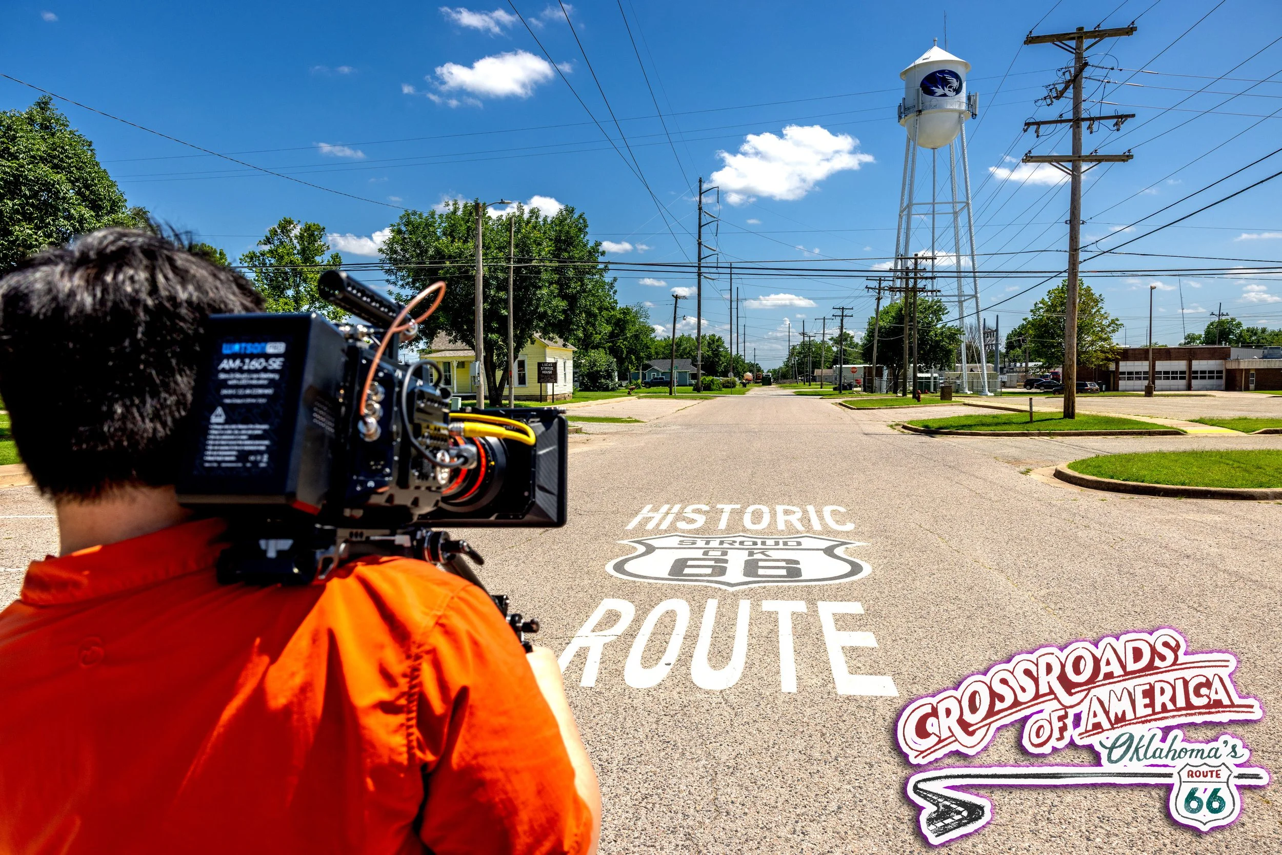 A person wearing an orange jacket holding a camera, standing on a road with Route 66 markings. There are trees, power lines, and a water tower in the background under a partly cloudy sky.