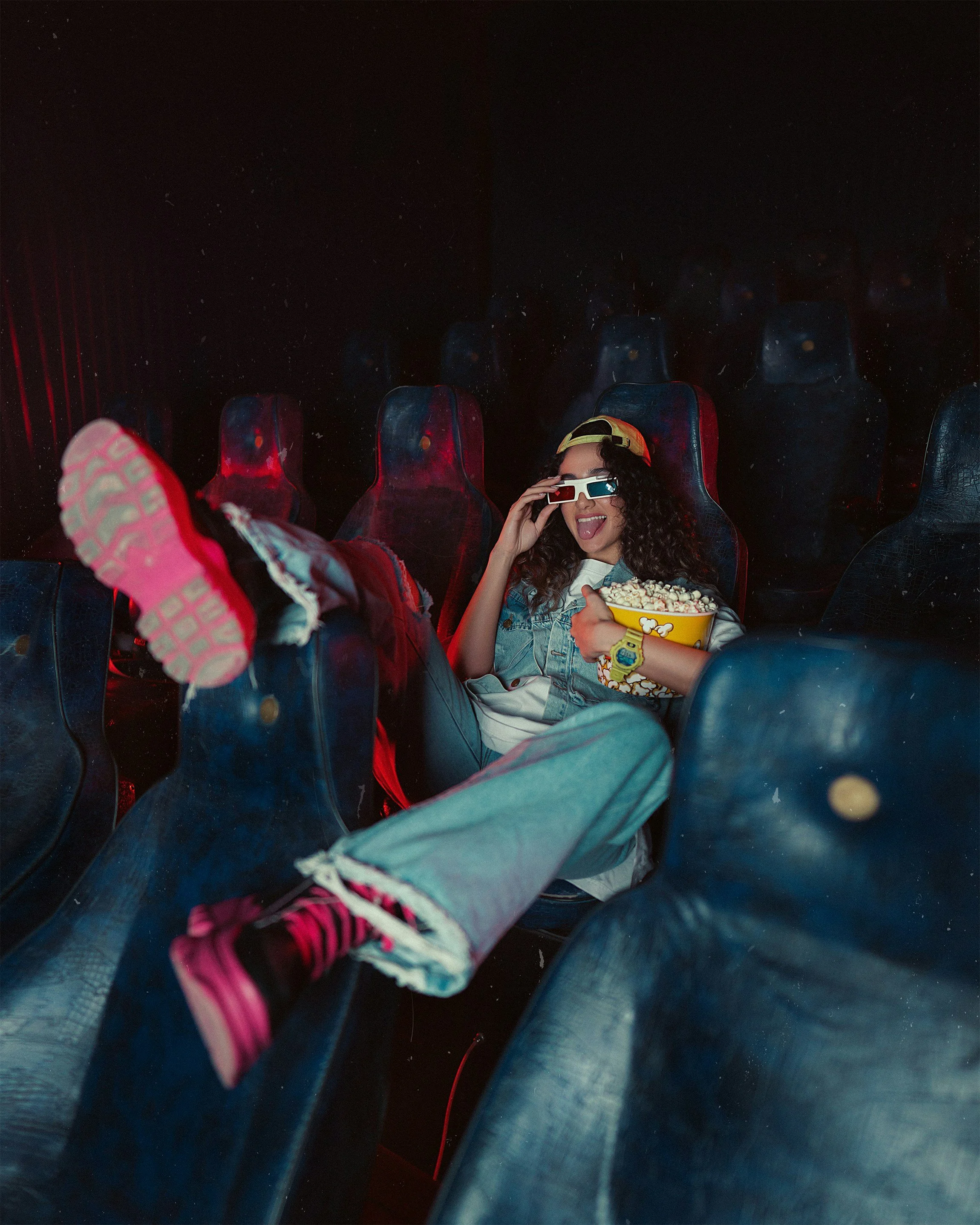 A young woman with curly hair wearing 3D glasses and a yellow cap, smiling and holding a large bucket of popcorn while sitting in a movie theater, with empty movie seats surrounding her.