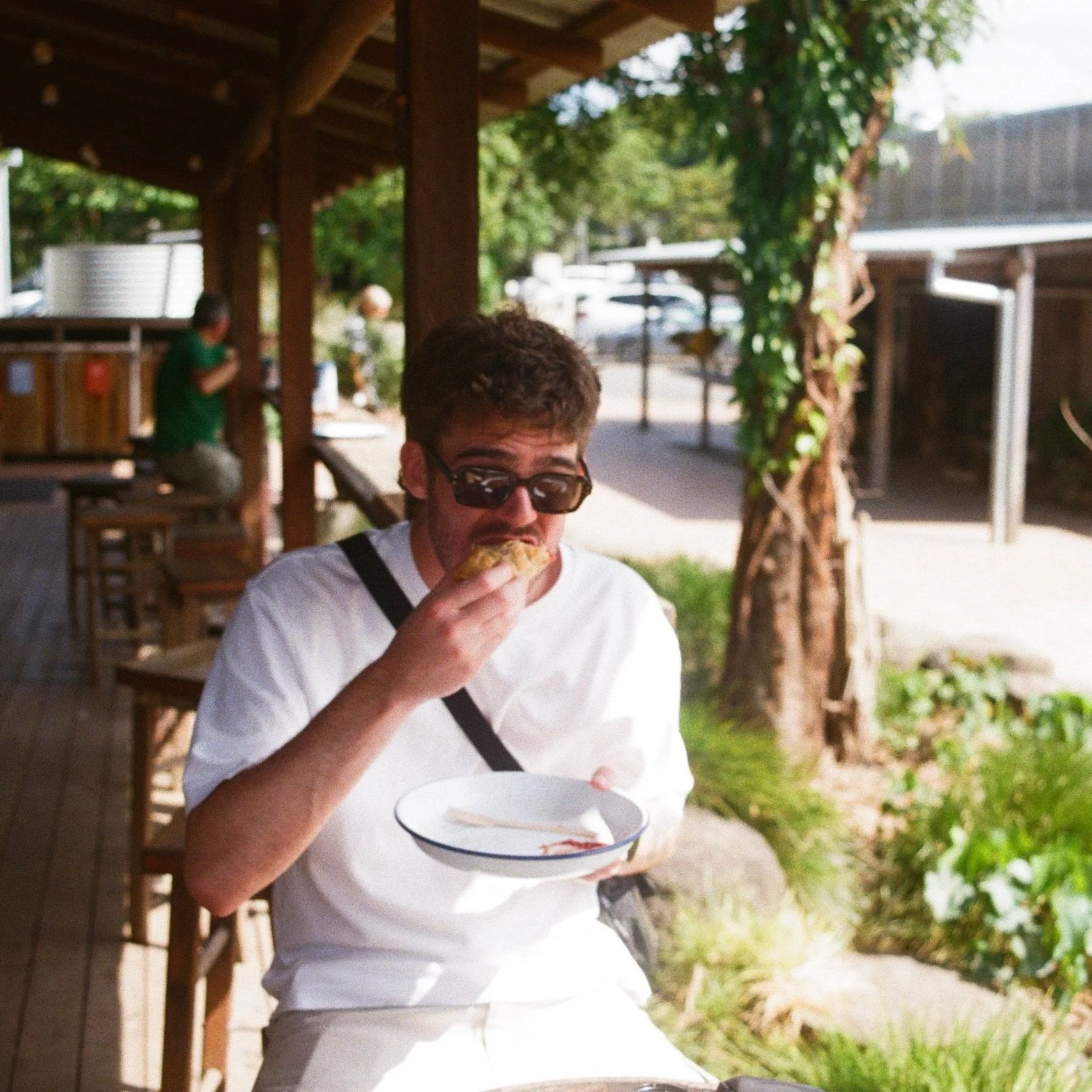 A young man with brown hair and sunglasses is eating a hamburger and holding a bowl in an outdoor setting near a wooden structure, trees, and greenery.