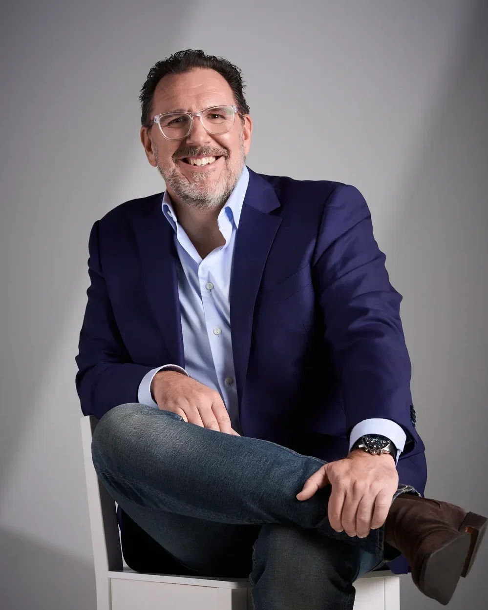 A smiling man with glasses in a blue blazer, white shirt, jeans, and brown shoes sitting on a white chair against a gray background.