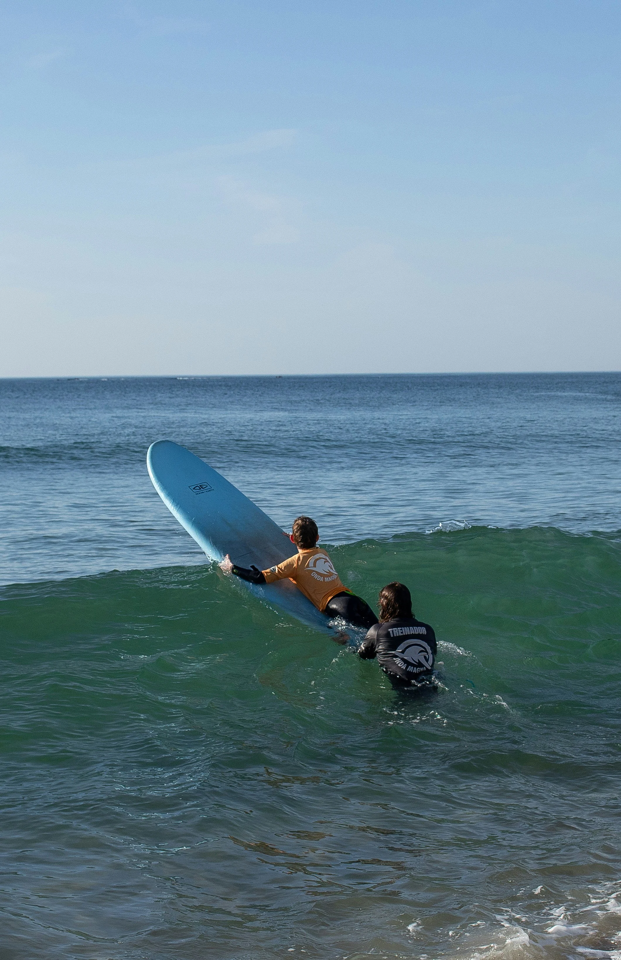 A person in a brown wetsuit holds a blue surfboard while another person in a black wetsuit assists them in the ocean near the shore during daytime, with the sky and sea visible in the background.