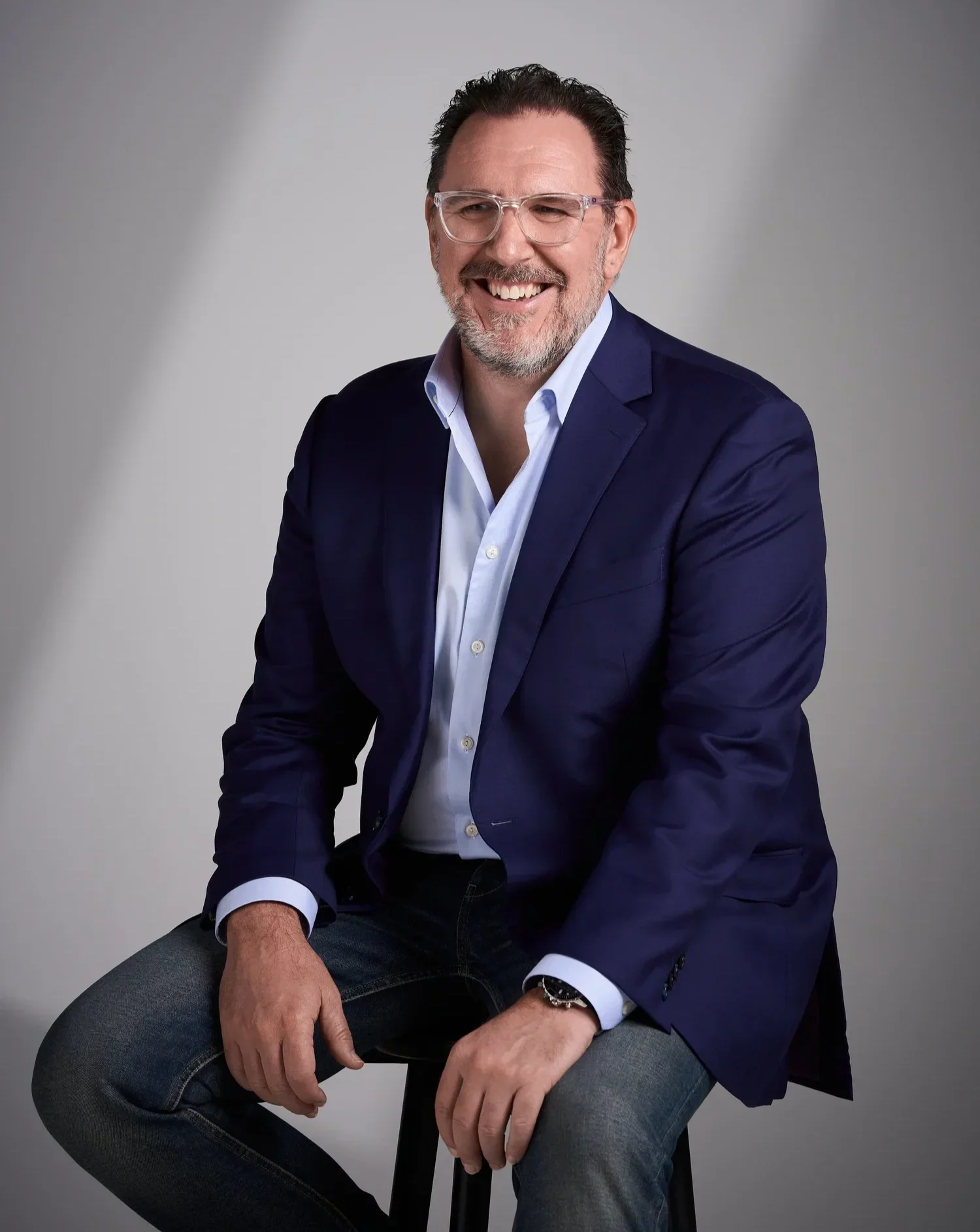 Man with dark hair, glasses, and a beard wearing a dark blue blazer and white shirt, sitting on a black stool against a plain gray background, smiling and looking to his left.