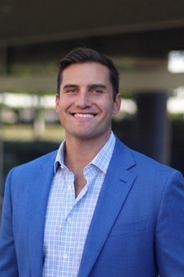 A man in a blue suit jacket and a light blue checkered shirt smiling outdoors near a bridge.