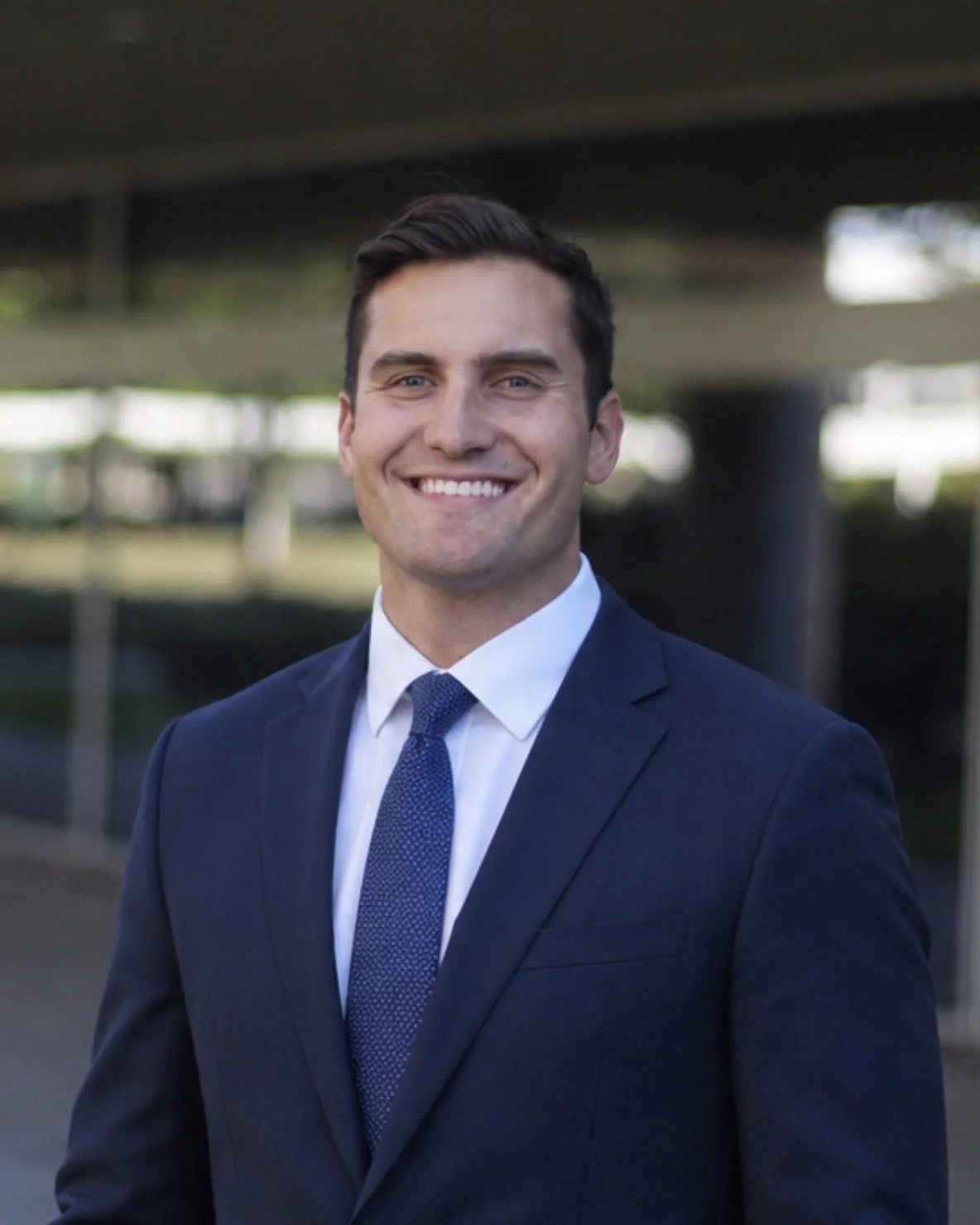 A young man in a dark suit, white shirt, and blue tie standing outdoors under a bridge, smiling at the camera.