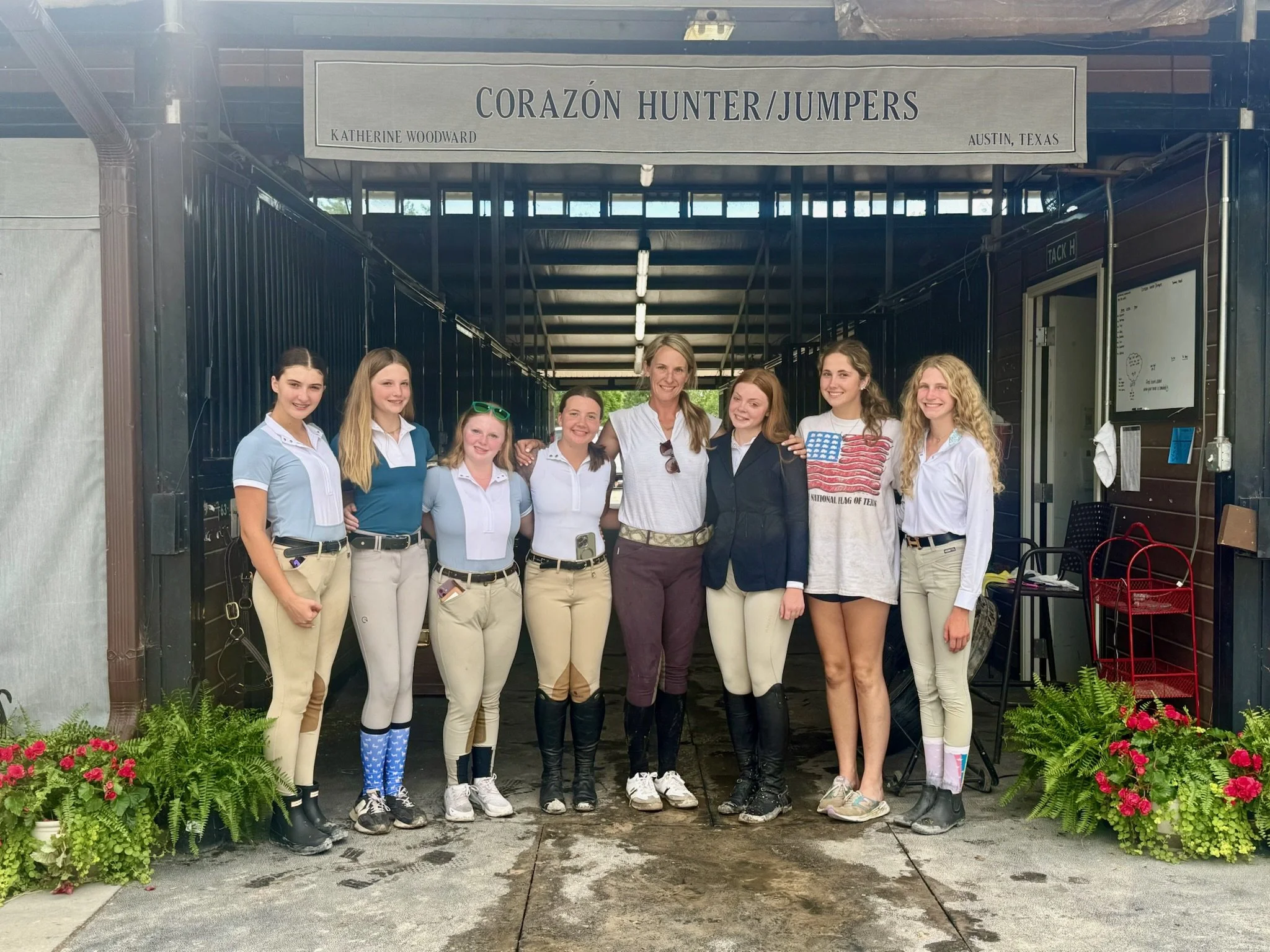 A group of nine young women and one woman standing together on a driveway in front of a building with a sign that reads 'Corazón Hunter/Jumpers, Katherine Woodward, Austin, Texas'. They are dressed in equestrian riding attire, and some are wearing boots, breeches, and polo shirts. There are potted plants with pink flowers at the sides of the group.