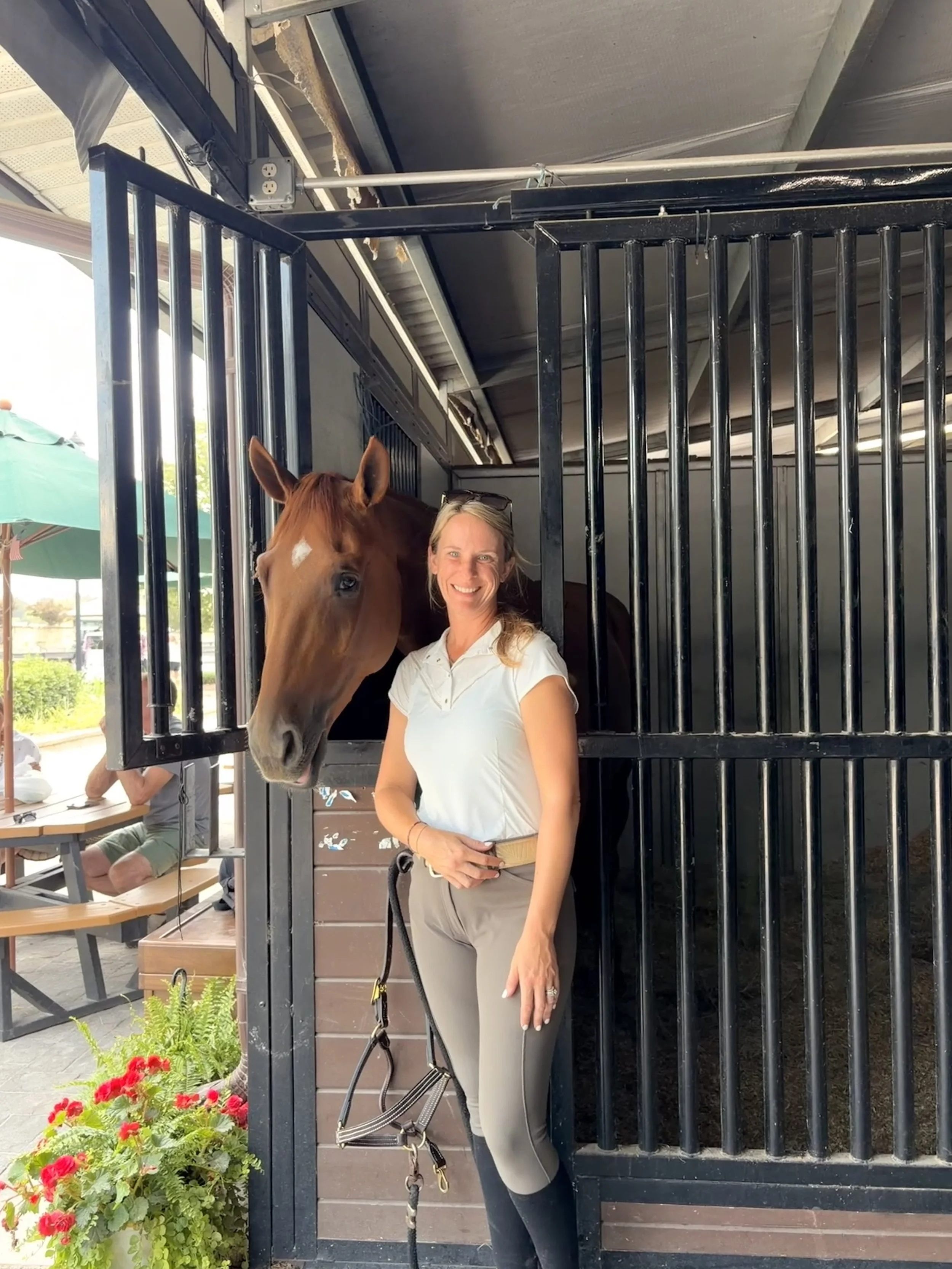 Woman standing next to a horse inside a black metal stall at an outdoor horse stable.