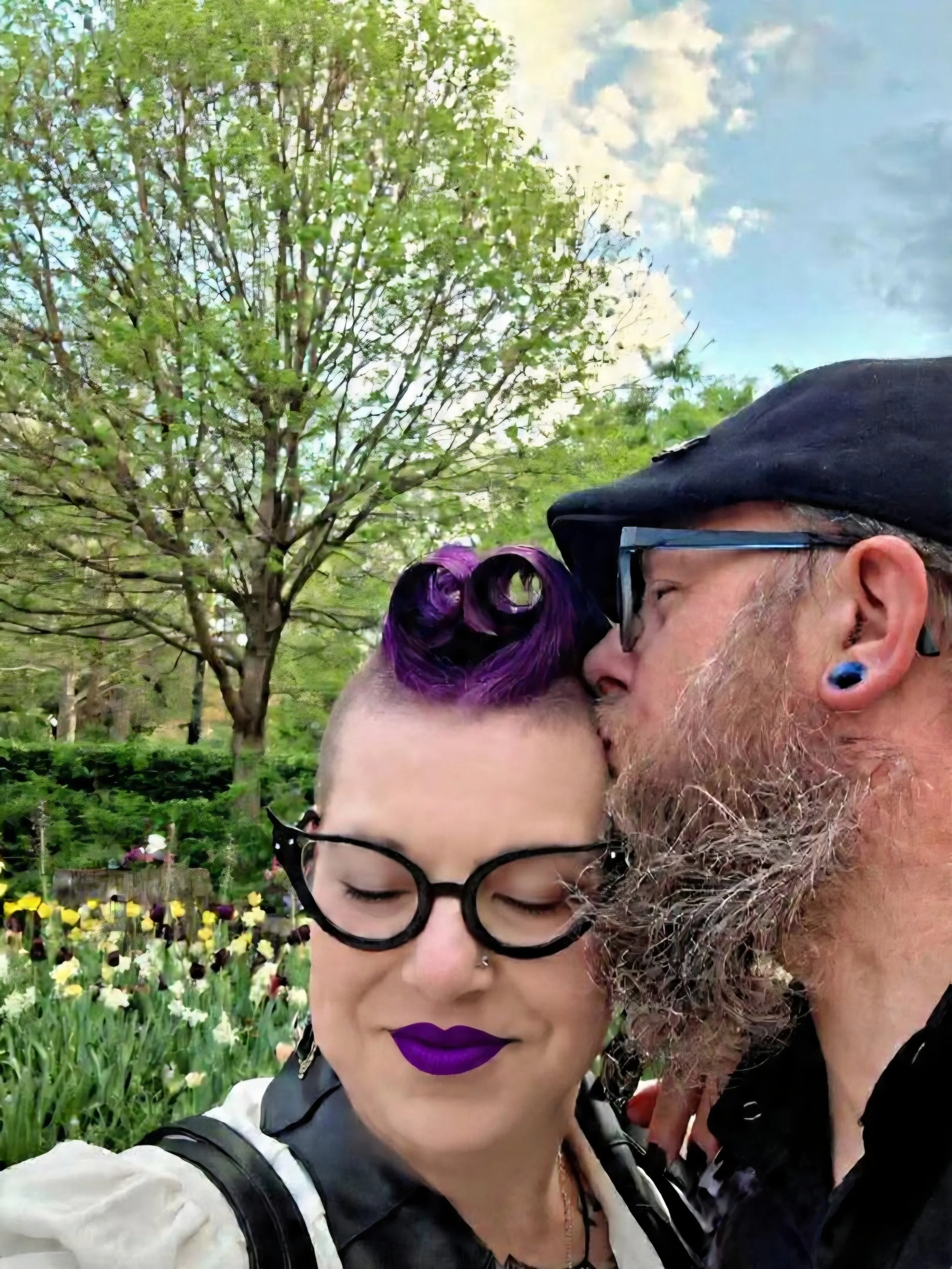A woman with black glasses and purple lipstick stands next to an older man with a beard and earrings, both with eyes closed, in a park with trees and flowers.