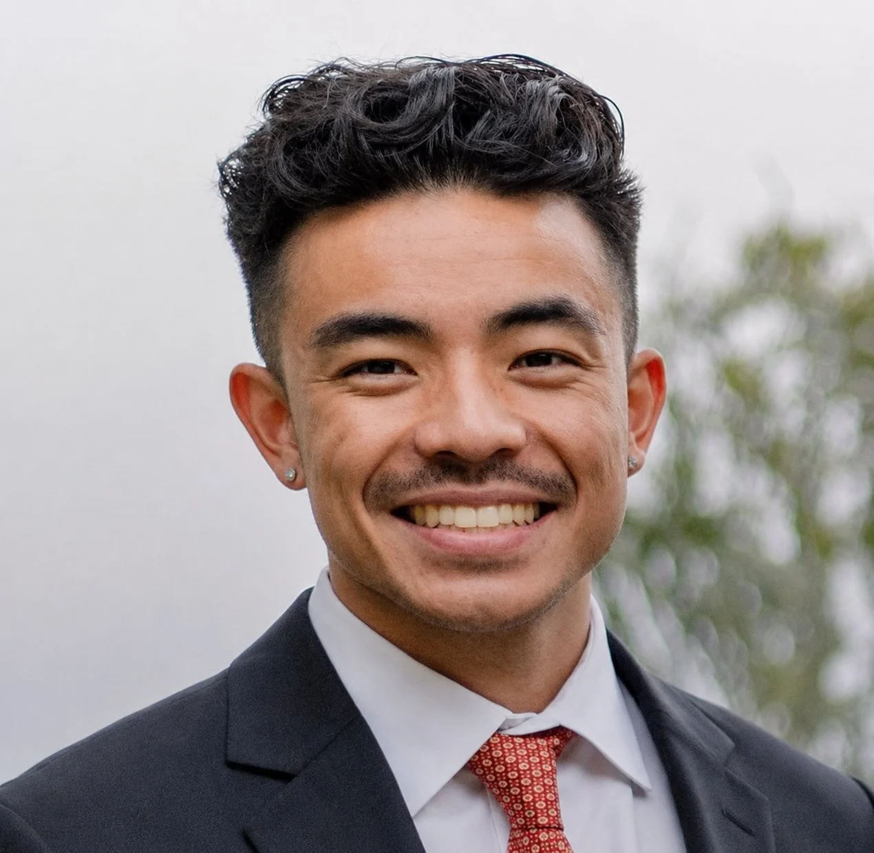 Smiling man in formal attire, including a suit and tie, outdoors with blurred trees in the background.