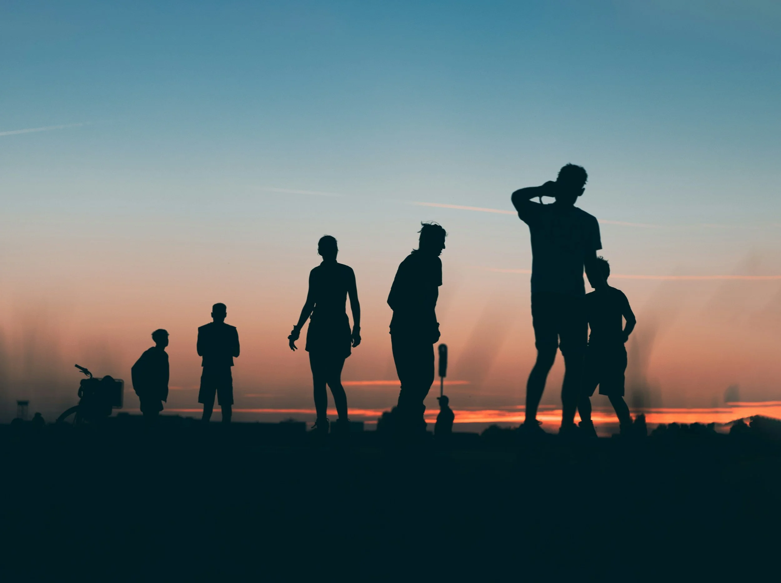 Silhouettes of people walking and standing on a beach during sunset, with a large sky in the background.