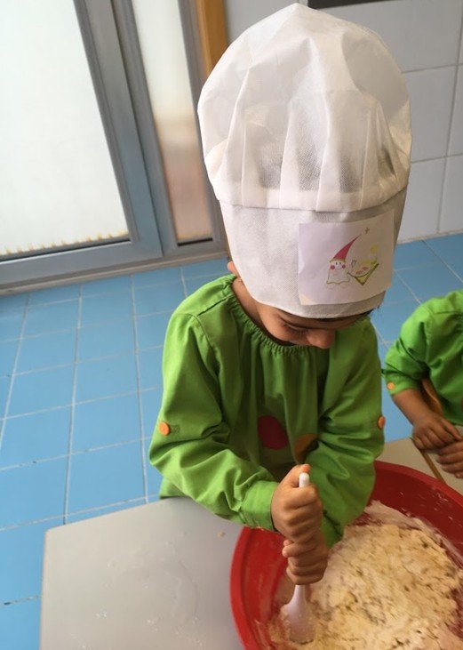 Criança com chapéu de cozinheiro, vestindo avental verde, mexendo uma mistura de massa em uma tigela vermelha.