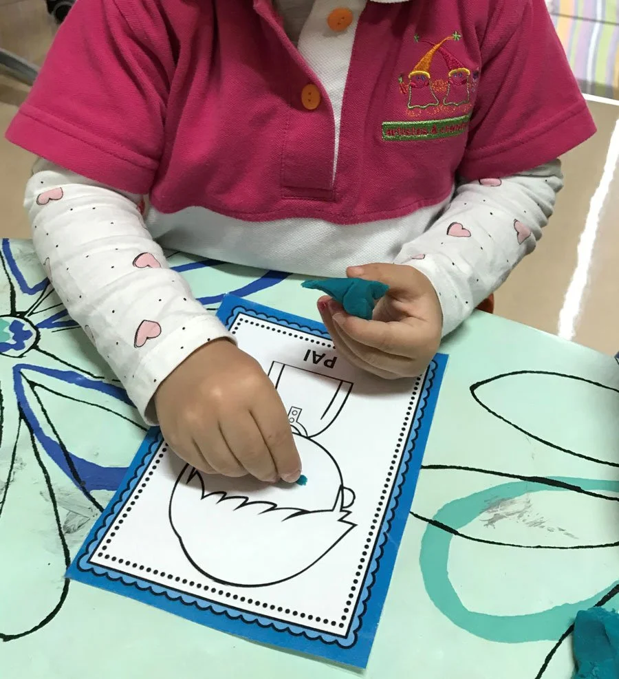 Criança colorindo uma folha de papel com desenho de uma ave em uma mesa com ilustrações de folhas, usando uma blusa de manga longa com corações e um uniforme rosa por cima. A criança está segurando um giz de cera azul.