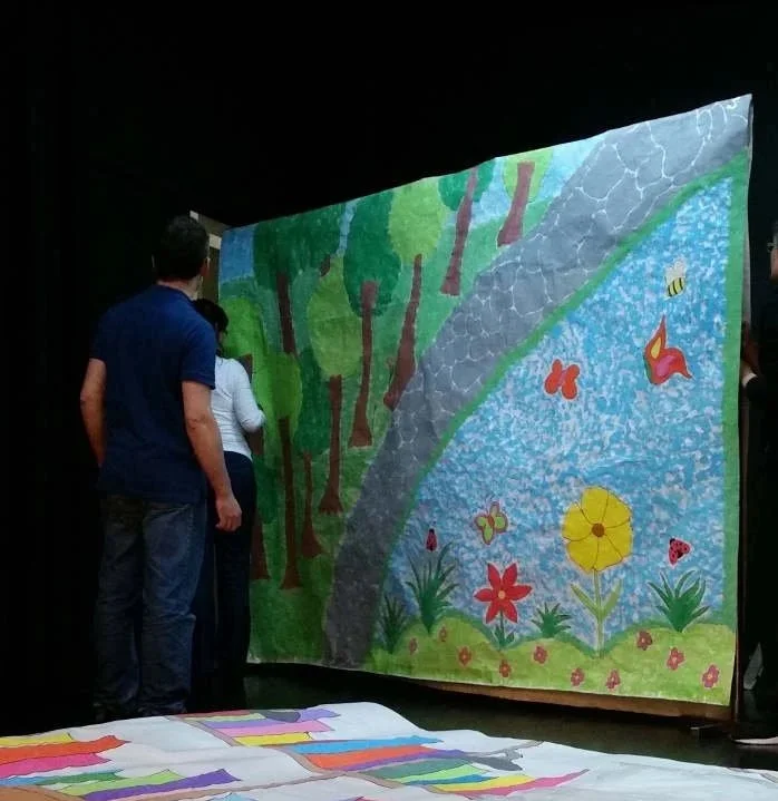 Pessoas observando uma grande pintura que retrata uma cena natural com árvores, rio, flores e insetos.