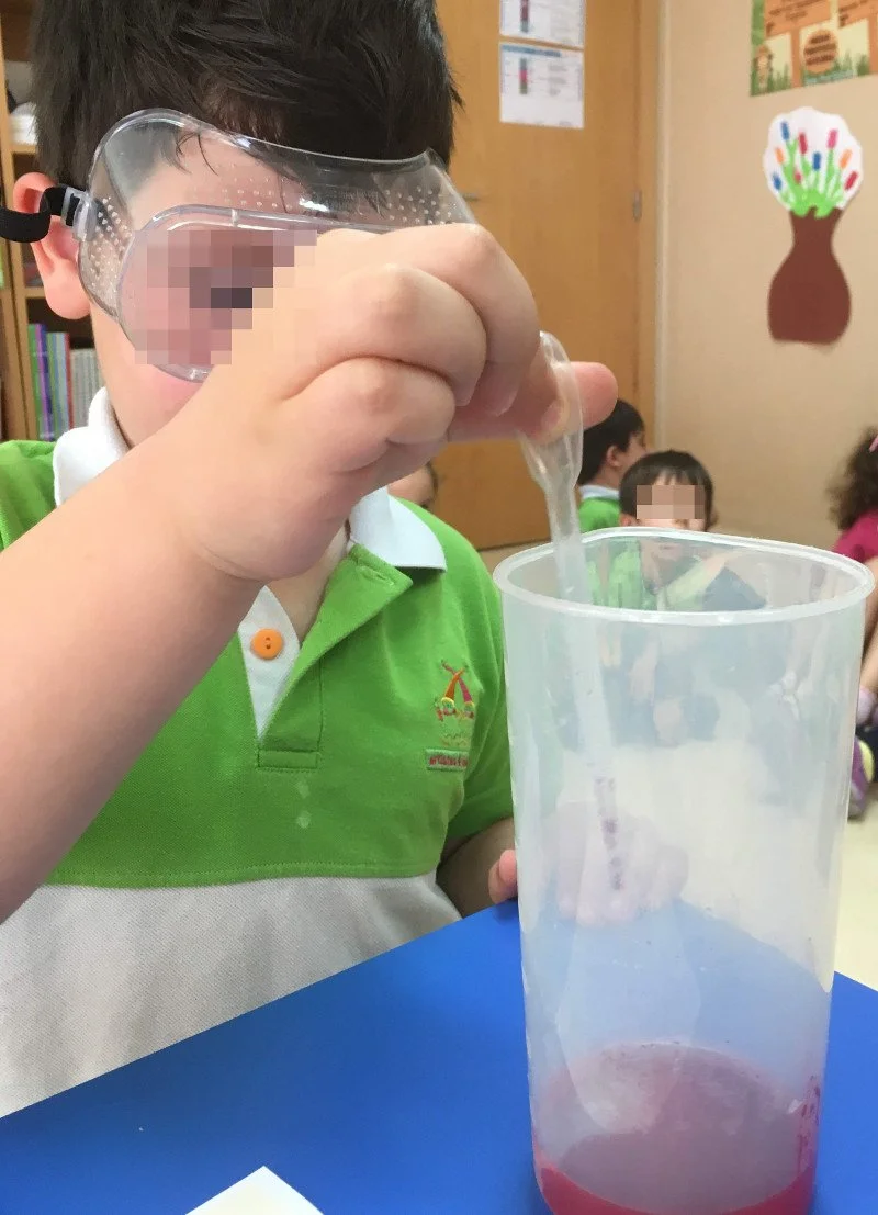 Criança com óculos de proteção coloridos, usando uma blusa verde e branca, adicionando líquido a um recipiente de plástico em uma sala de aula.