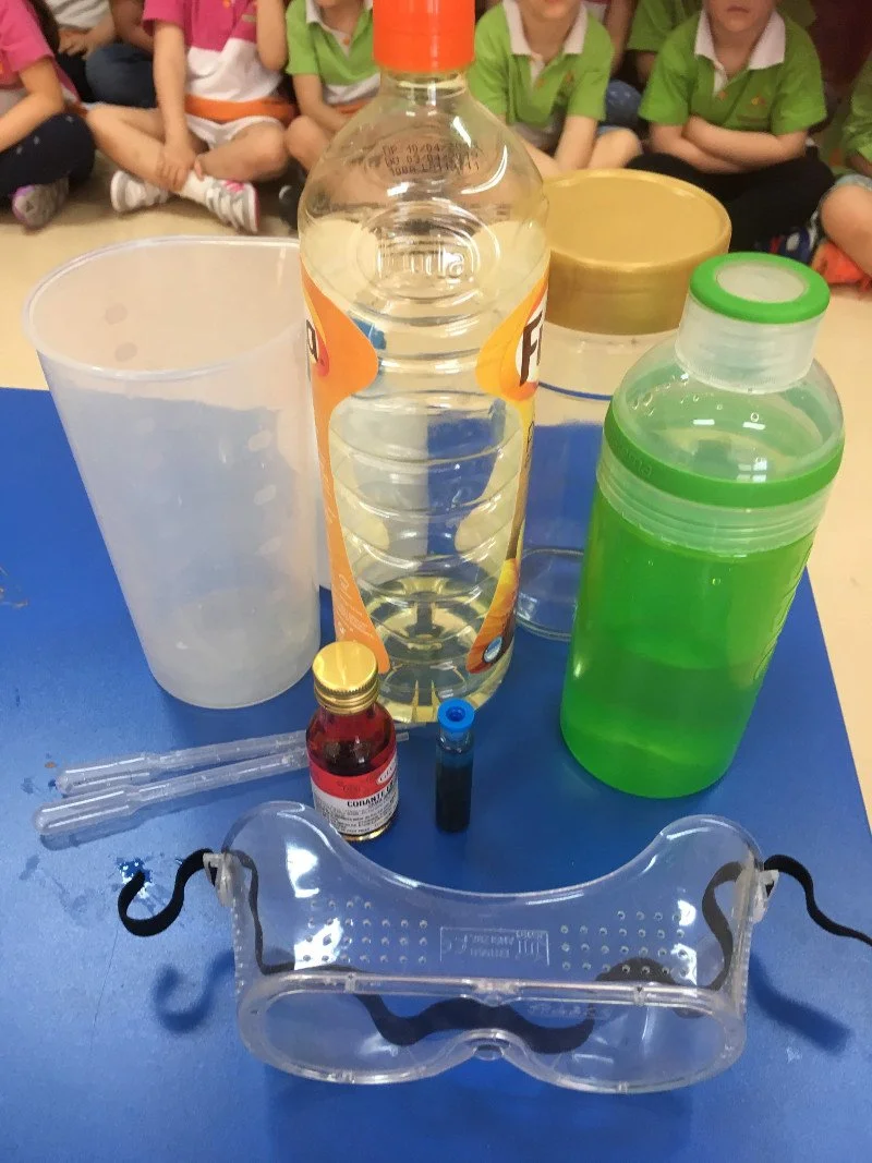 Itens para experiência científica: uma garrafa com água, uma garrafinha verde, uma garrafinha de refrigerante, uma tampa dourada, uma pequena garrafinha de remédio, duas pipetas, uma máscara de proteção transparente, e um grupo de crianças sentadas a