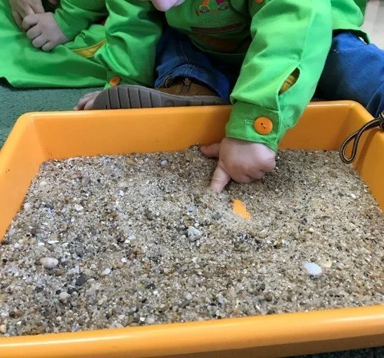 Criança brincando com caixa de areia em ambiente interno, usando jaqueta verde e calça jeans, apontando para a areia com o dedo.