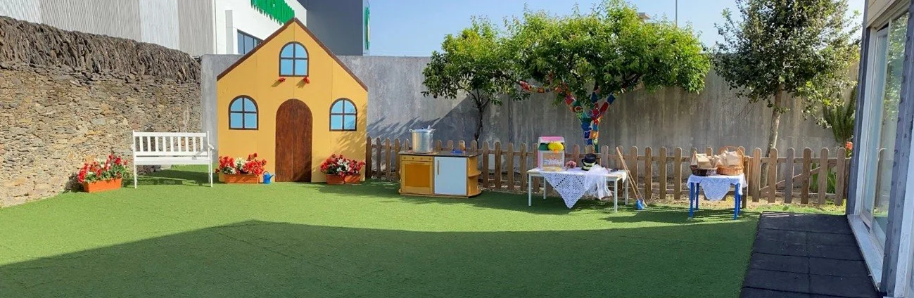 Arte de uma casinha de brinquedo decorada com flores, uma mesa com doces e um balde no quintal de uma casa, com árvores e uma cerca de madeira ao fundo.