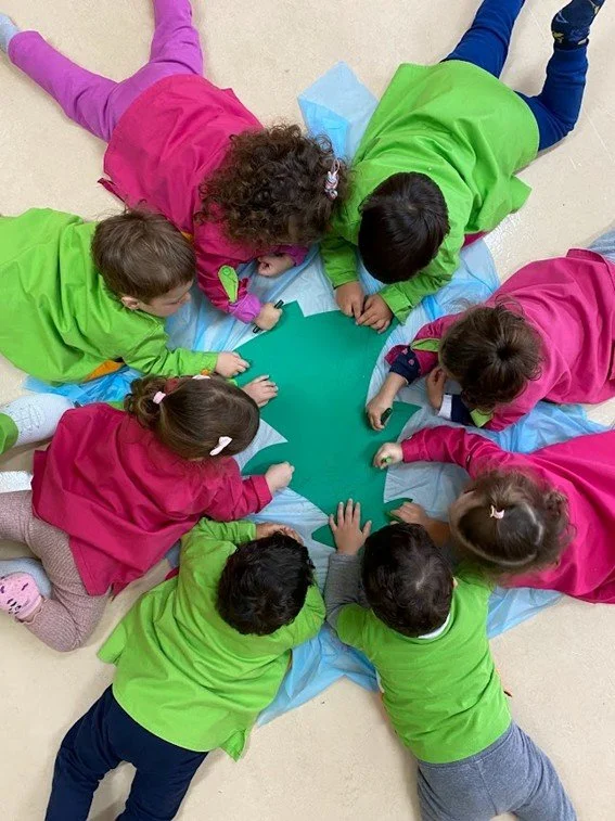 Crianças usando jalecos coloridos, algumas verdes e outras rosas, estão deitadas em círculo no chão ao redor de um coração de papel verde, brincando e interagindo.