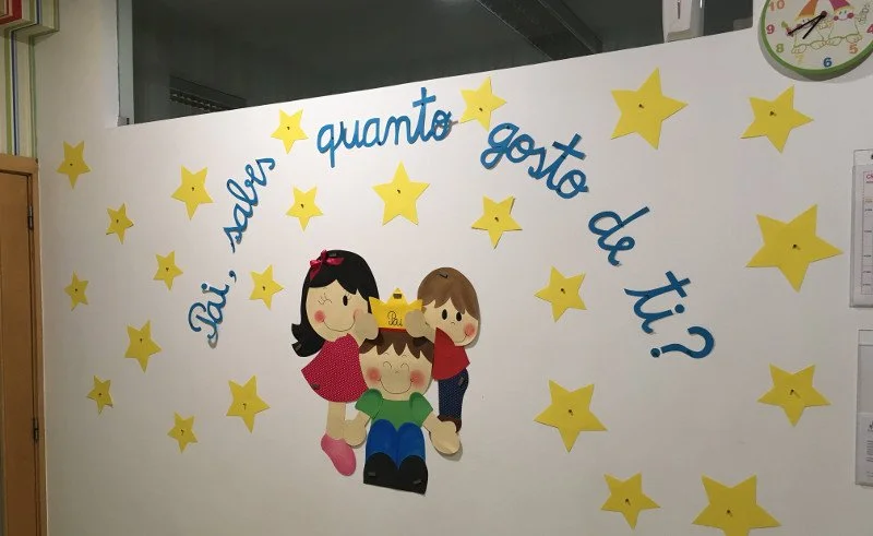 Parede decorada com estrelas amarelas e personagens de desenho infantil, incluindo uma menina com cabelo preto, uma menina com cabelo castanho e um menino com cabelo castanho claro, todos sorrindo. Texto em azul na parede que diz 'Até aqui, quanto go