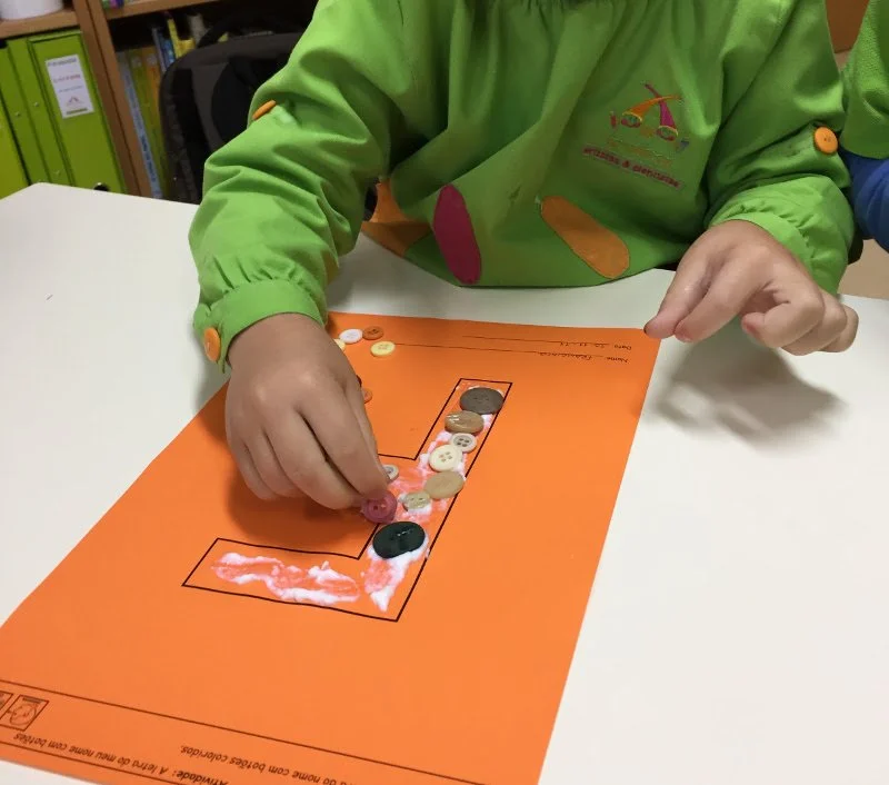 Criança colocando botões em um tabuleiro em forma de letra 'L' com um fundo laranja, em uma mesa branca, em ambiente escolar.
