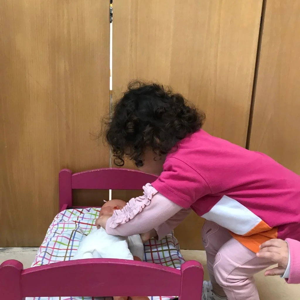 Criança ajudando uma bebê a virar na cama em uma cama de madeira rosa, em um ambiente interno com portas de madeira ao fundo.