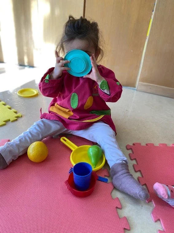 Criança sentada em um tapete de EVA rosa, segurando um copo azul na frente do rosto, com brinquedos ao redor, incluindo uma bola de limão, uma colher de chá amarela, um copo azul em uma bandeja vermelha, uma berinjela de brinquedo, e um sapato cinza.