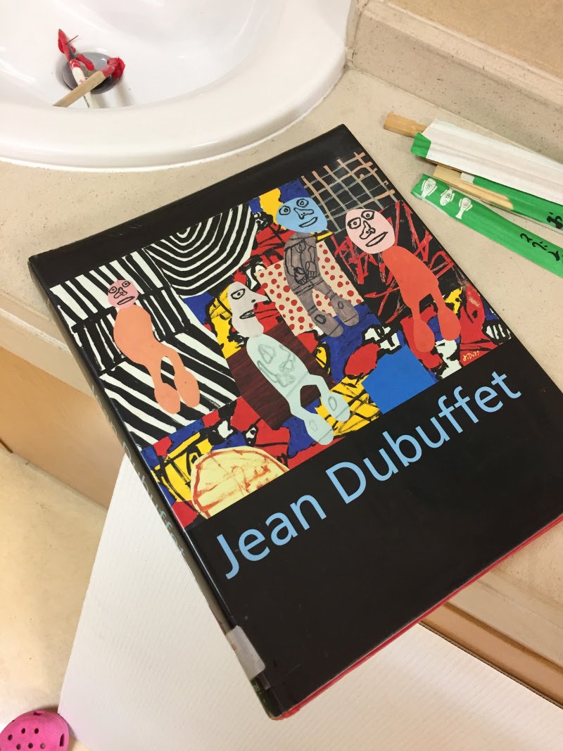 Livro de arte de Jean Dubuffet com capa colorida, incluindo figuras abstratas, linhas e formas diversas, repousando em uma bancada de banheiro ao lado de objetos de papelaria.