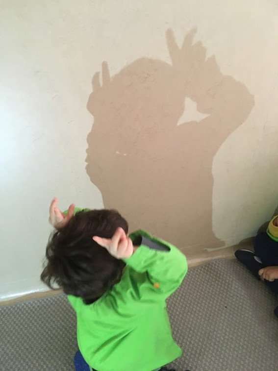 Criança de cabelo escuro vestida com blusa verde, olhando para a sombra na parede, que se assemelha a uma silhueta com chifres, criando a ilusão de um personagem com chifres na sombra.