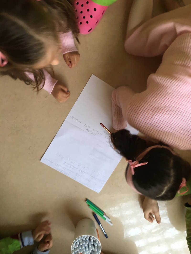 Crianças brincando e escrevendo uma carta ou poema em uma folha de papel sobre o chão, com canetas ao lado.
