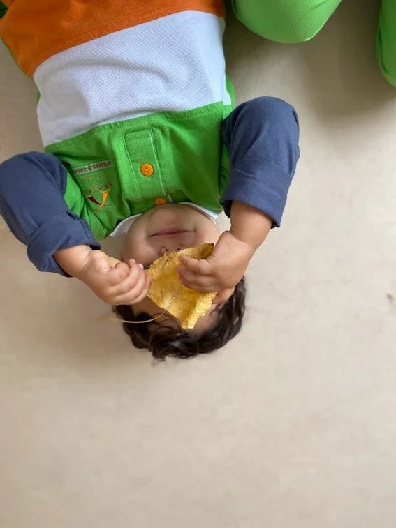 Criança deitada no chão, segurando uma fatia de biscoito, sorrindo e usando roupas coloridas, incluindo blusão azul e calça verde.