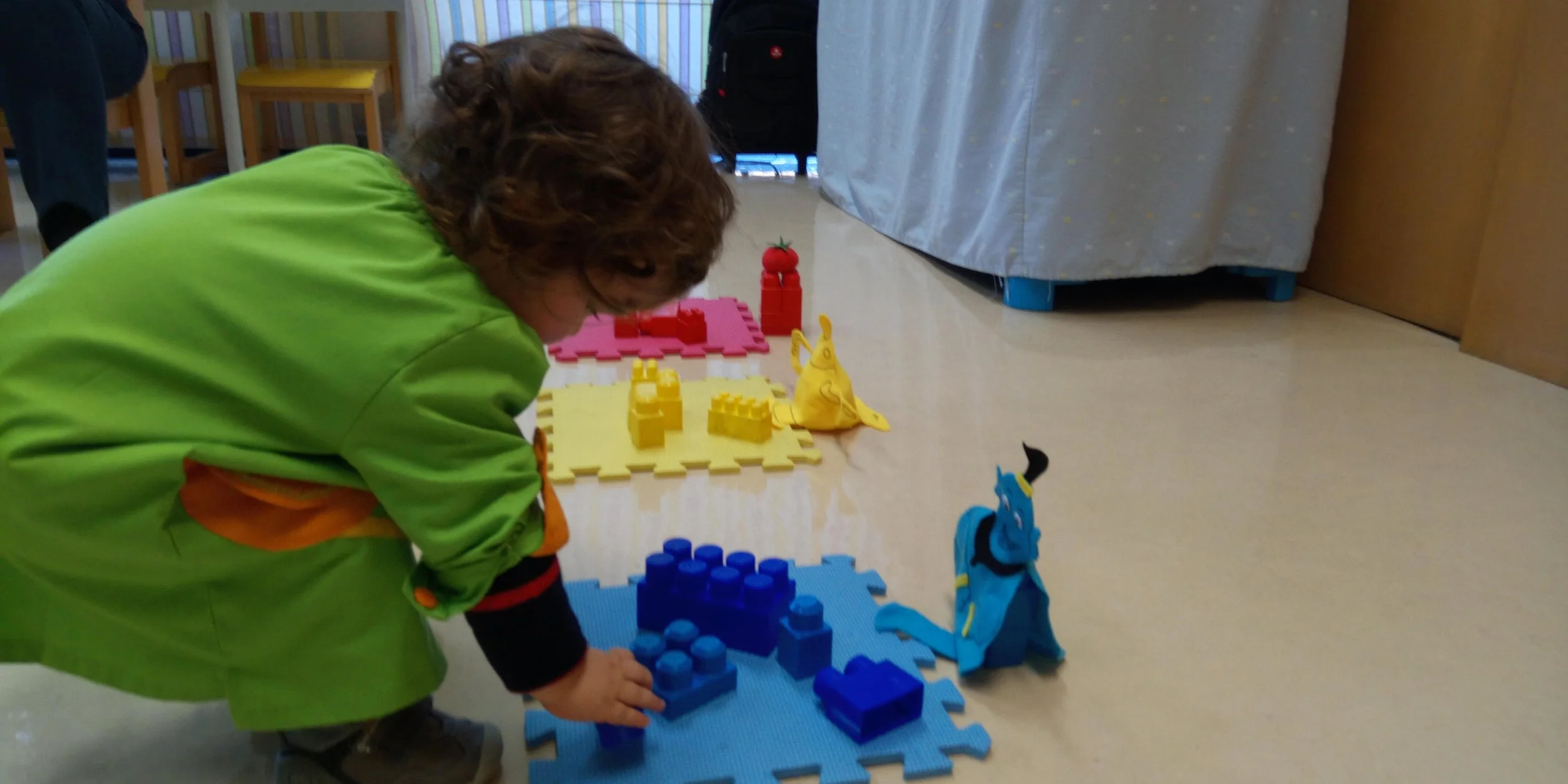 Criança brincando com blocos de montar em um ambiente interno, com brinquedos e cadeiras ao fundo.