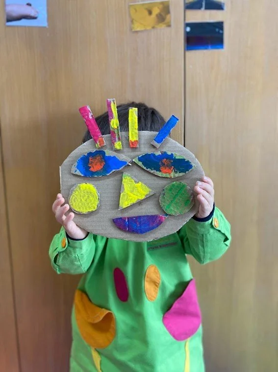 Criança segurando uma máscara colorida feita de papelão, com detalhes em cores vibrantes, em uma sala de aula.