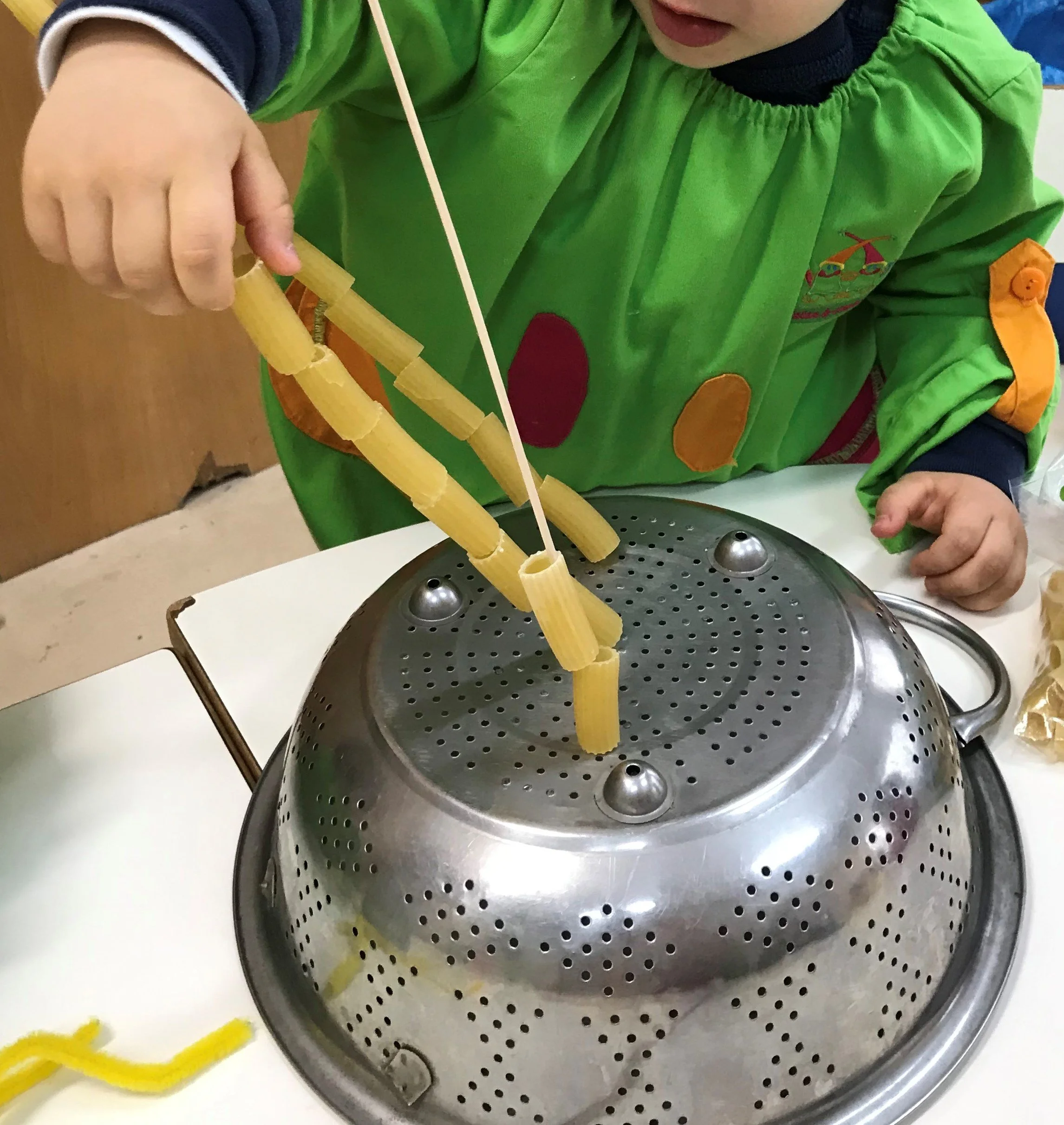 Criança usando avental verde colorido, realizando uma atividade com tubos de macarrão rígido e uma panela de cozimento.