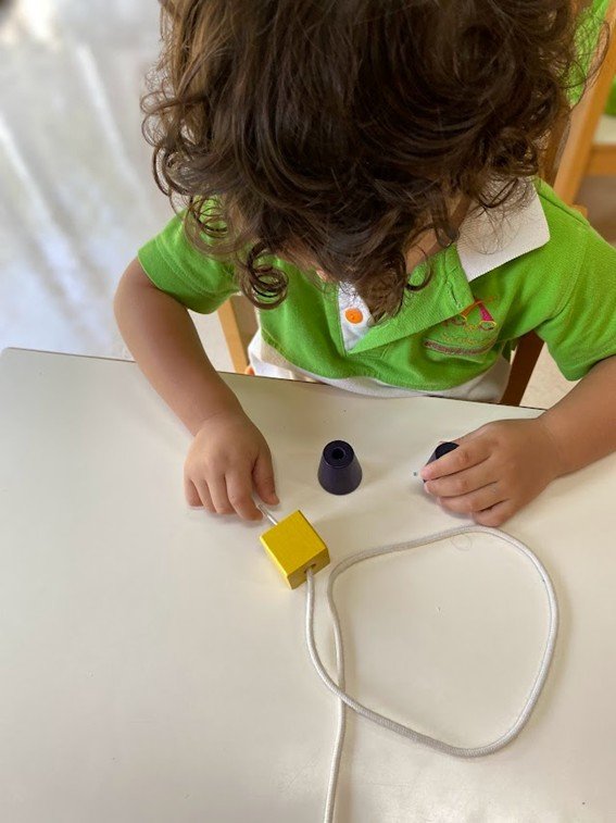 Criança com cabelo cacheado usando uniforme verde e branco, sentado à mesa, segurando uma peça preta de brinquedo ou ferramenta, com outros objetos como uma peça preta cônica e uma caixa amarela ao lado.