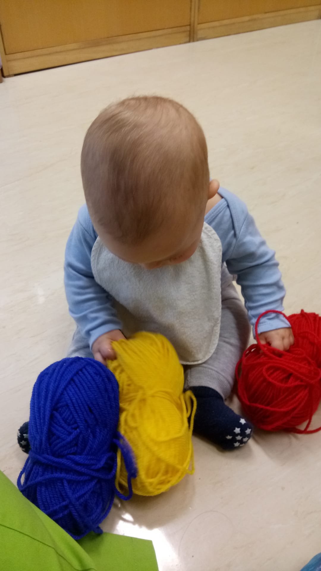 Criança sentada no chão segurando fios de lã azul, amarelo e vermelho, com teias de lã próximas.