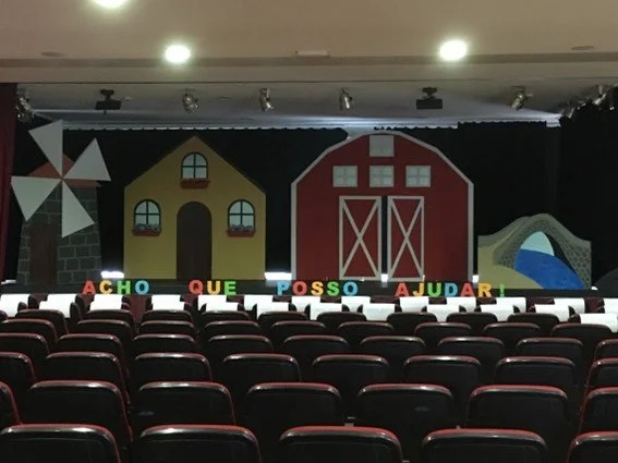 Palco de teatro com cenário de fazenda, incluindo uma casa amarela, um celeiro vermelho, uma roda de moinho e uma cerca, com cadeiras em primeiro plano.