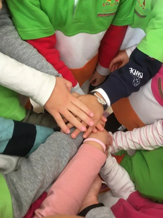 Mãos de crianças sobrepostas em um gesto de união, usando roupas variadas e coloridas.