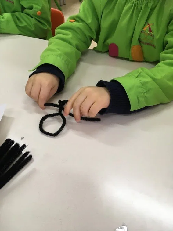 Criança vestida com jaqueta verde brincando com uma barra de cabelo de elástico preto com forma de laço em uma mesa branca.