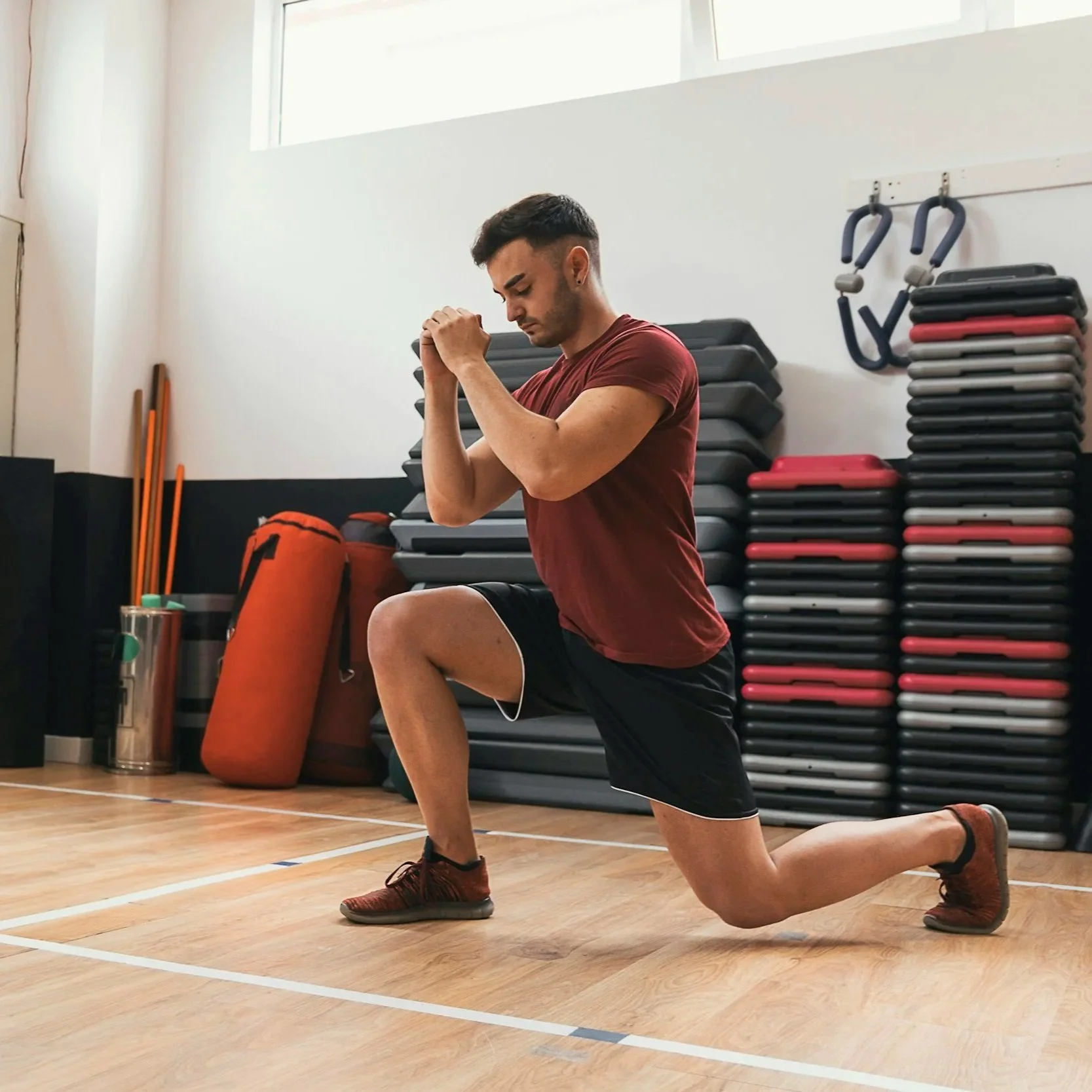 A man in athletic clothing performing a lunge exercise in a gym, with fitness equipment and stacked steps in the background.