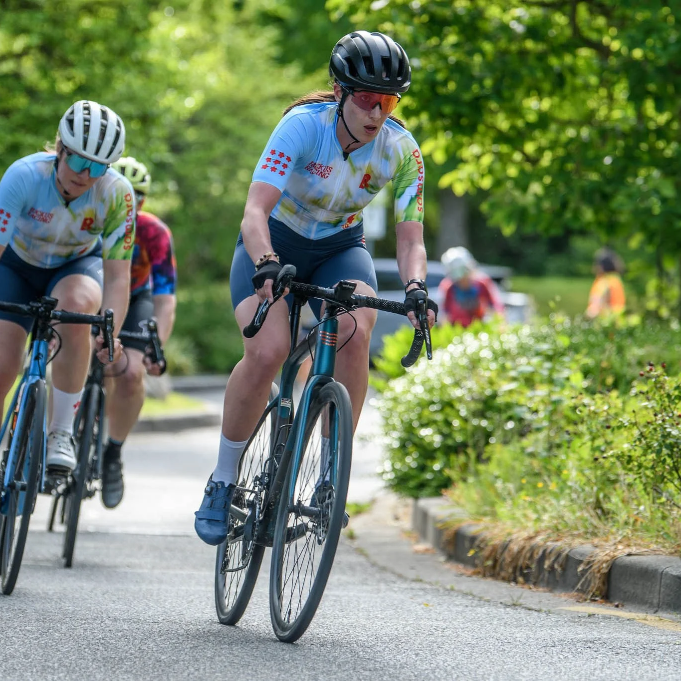 Two women riding bicycles on a paved road in a green, wooded area, wearing helmets and sports jerseys.