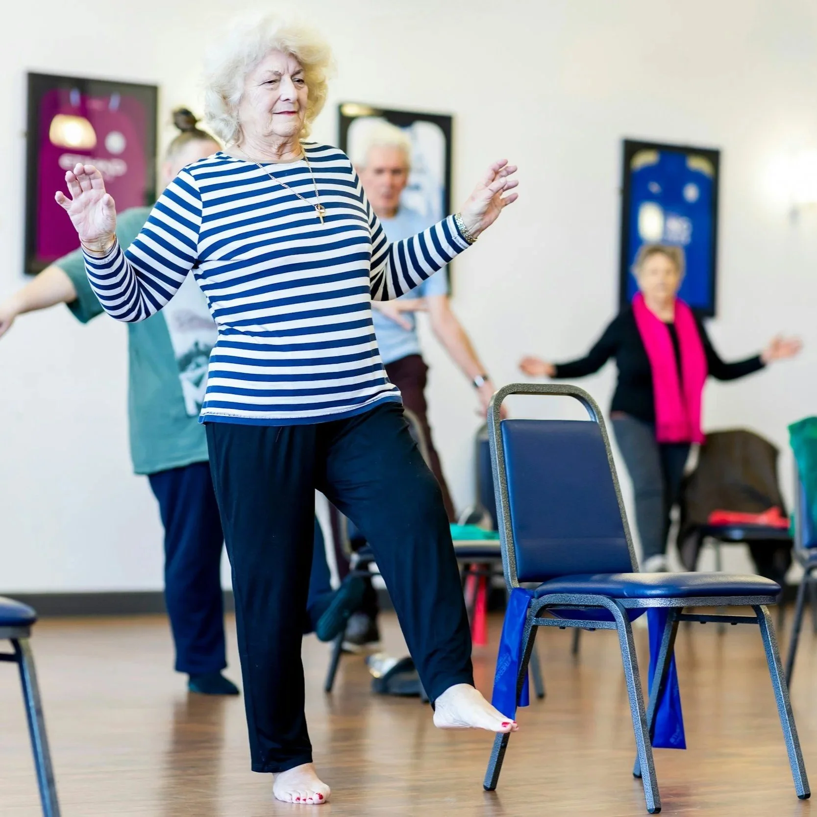 An elderly woman in a striped shirt practicing a balance exercise by lifting her leg onto a chair in a room with other women and chairs, possibly during a fitness or wellness class.