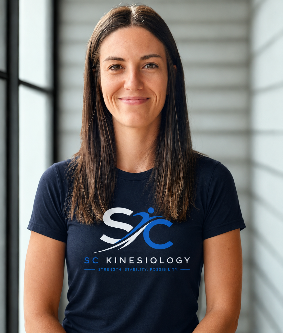 A woman with long brown hair, smiling, standing in front of a windowed wall, wearing a navy blue t-shirt with the logo and text "SC Kinesiology" and the tagline "Strength. Stability. Possibility."