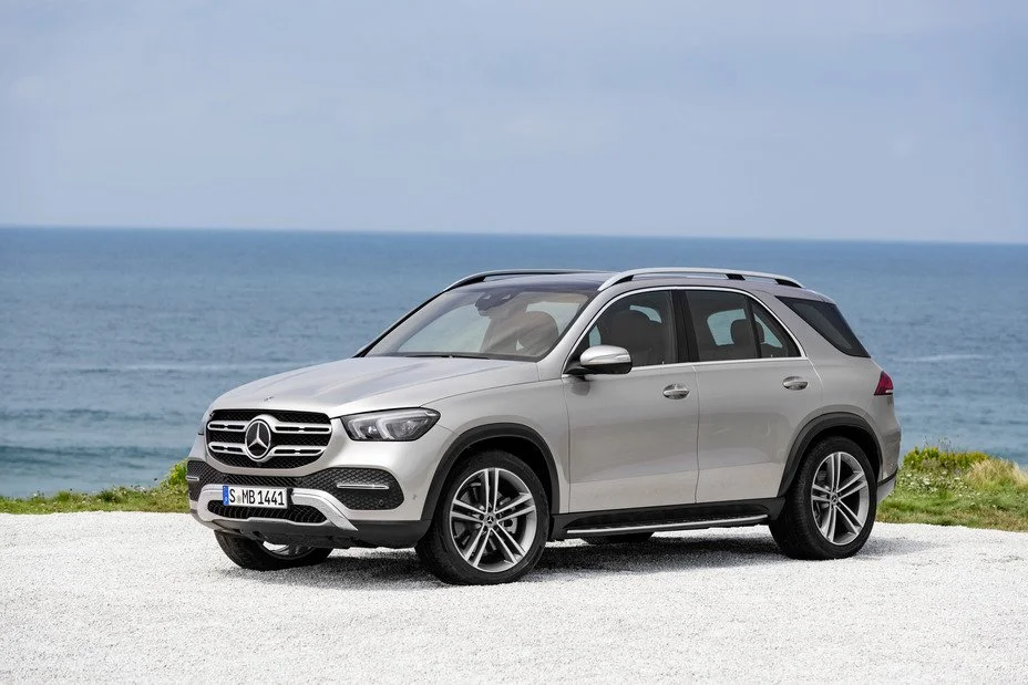 A silver Mercedes-Benz SUV parked on a gravel surface near the ocean, with a cloudy sky in the background.