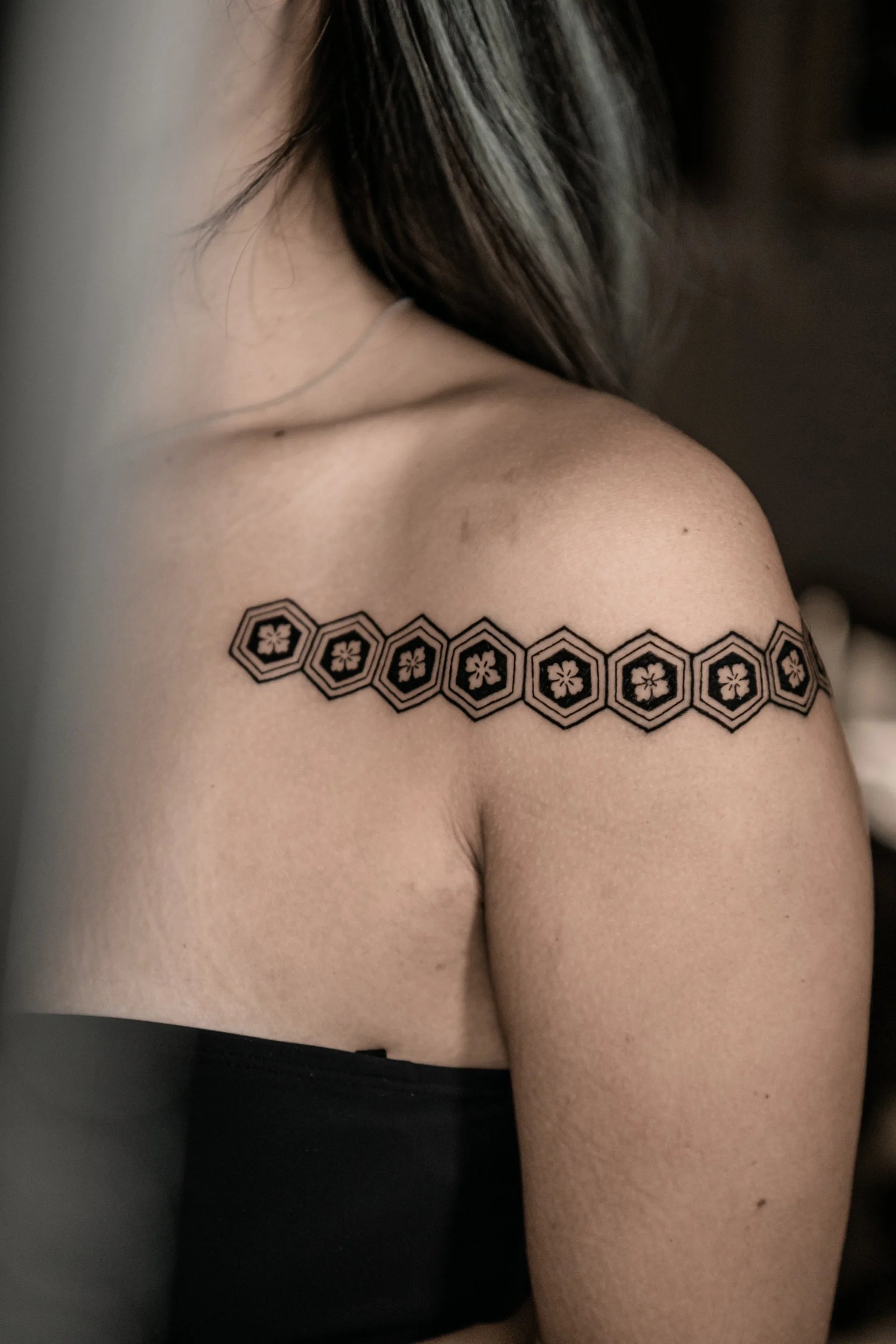 Close-up of a woman's shoulder and upper arm, featuring a black tattoo of interconnected hexagons with floral patterns inside each hexagon.