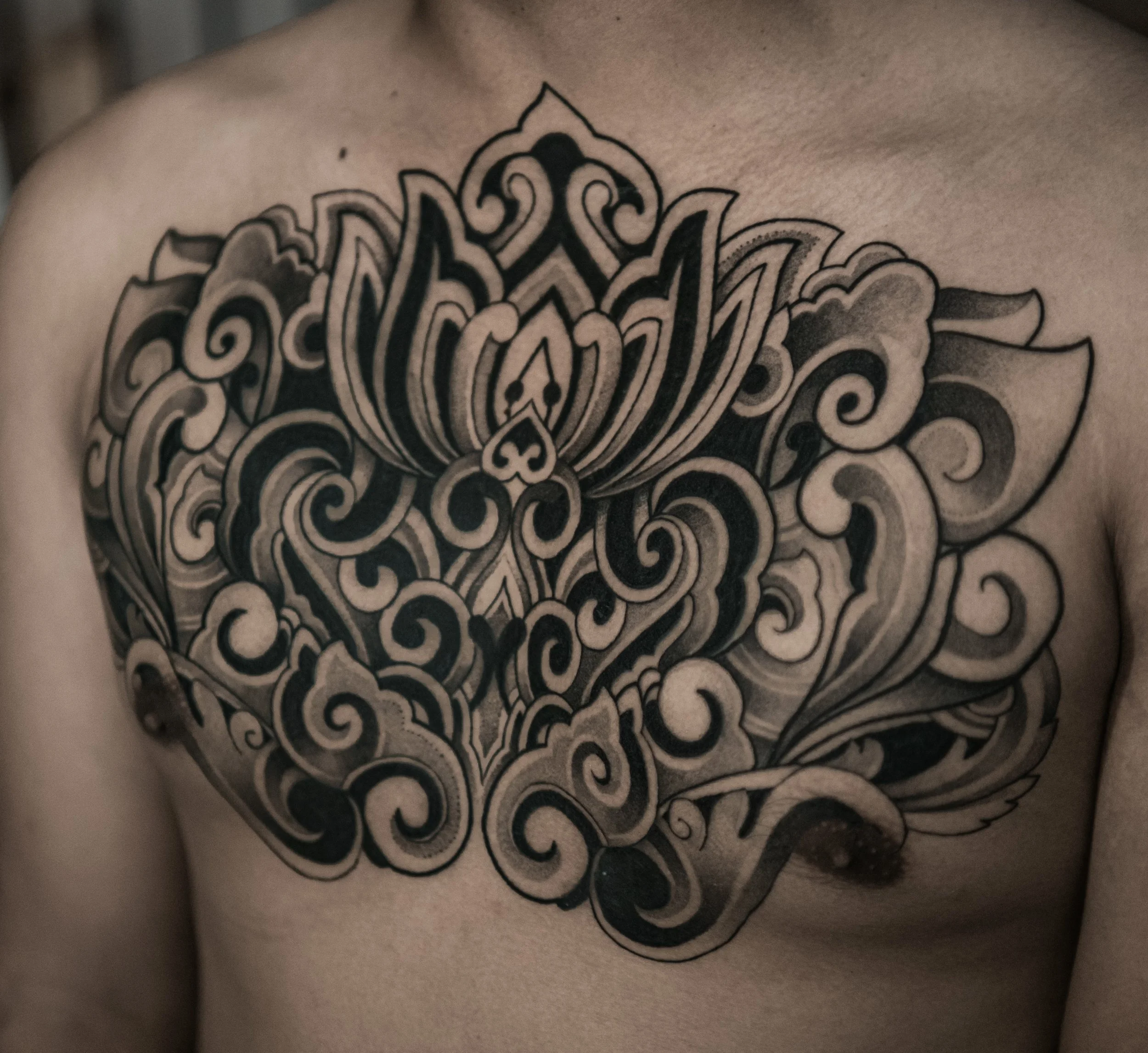 Close-up of a tattoo on a person's chest, featuring intricate black and gray decorative patterns resembling a mandala or floral design.