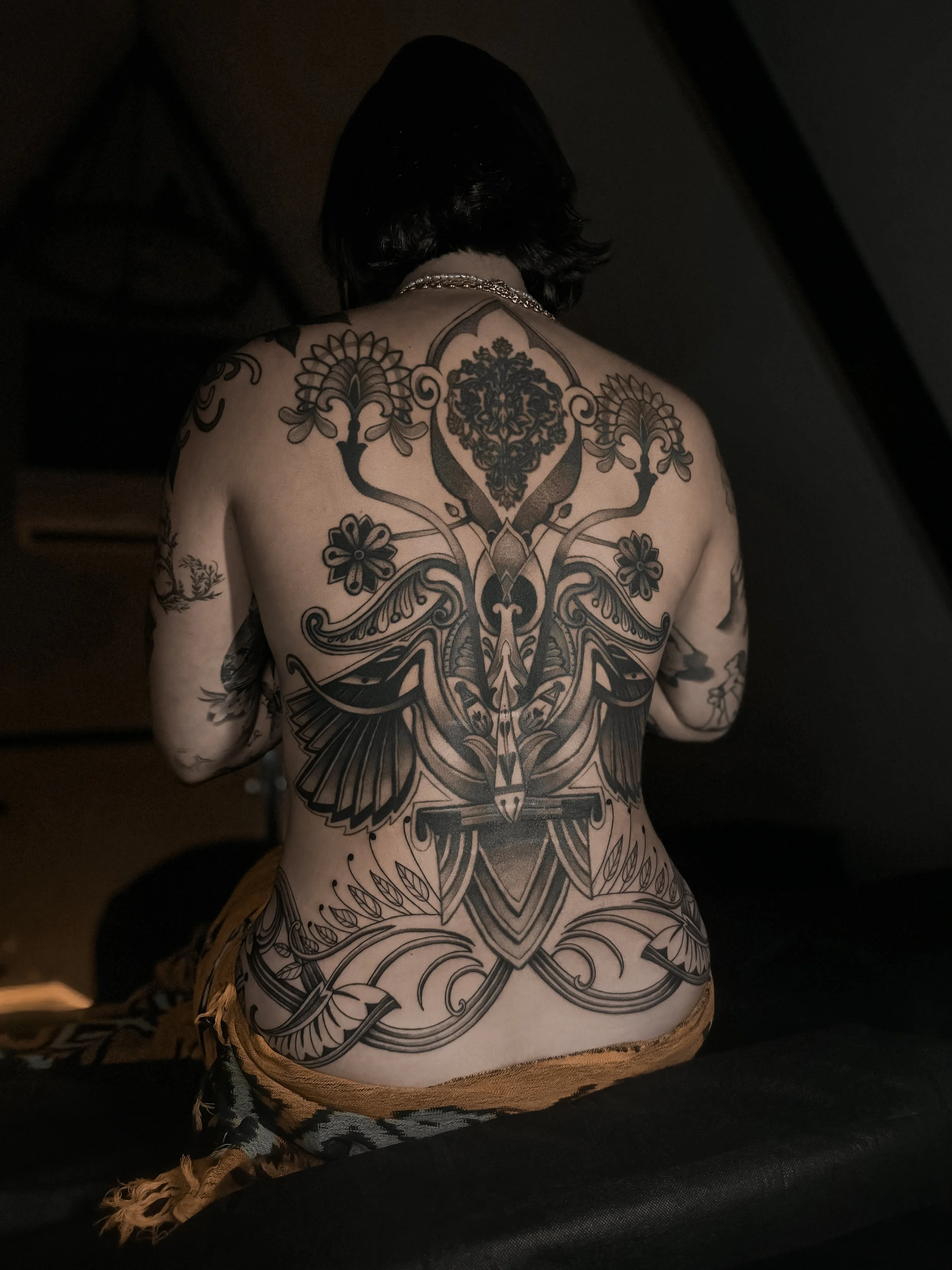 A person with black hair and a shiny chain necklace, showing their back and shoulders, with intricate black tattoo designs covering their back and arms, including floral and geometric patterns.
